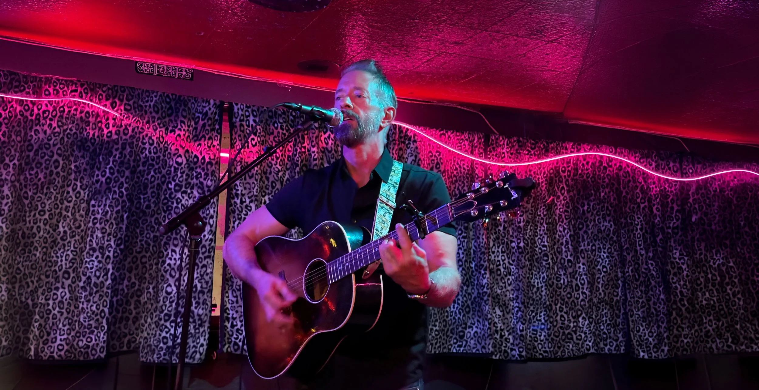 A man with a beard and short hair playing an acoustic guitar on a small stage in a dive bar with a neon pink light and leopard print curtain background.