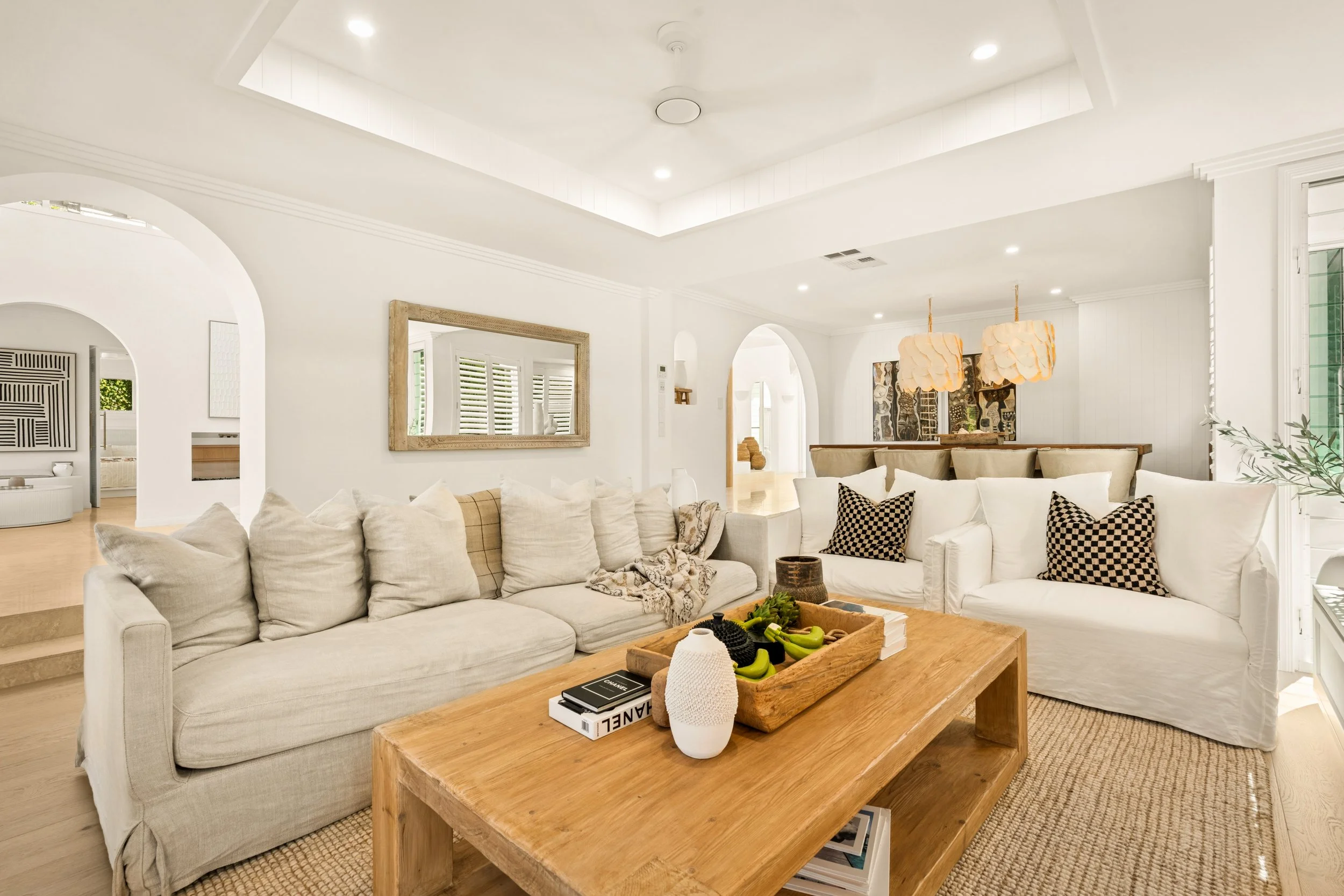 A bright, white living room with two large white sofas, decorative pillows, and a wooden coffee table with books and decorative items, featuring recessed ceiling lights and a large picture mirror on the wall.