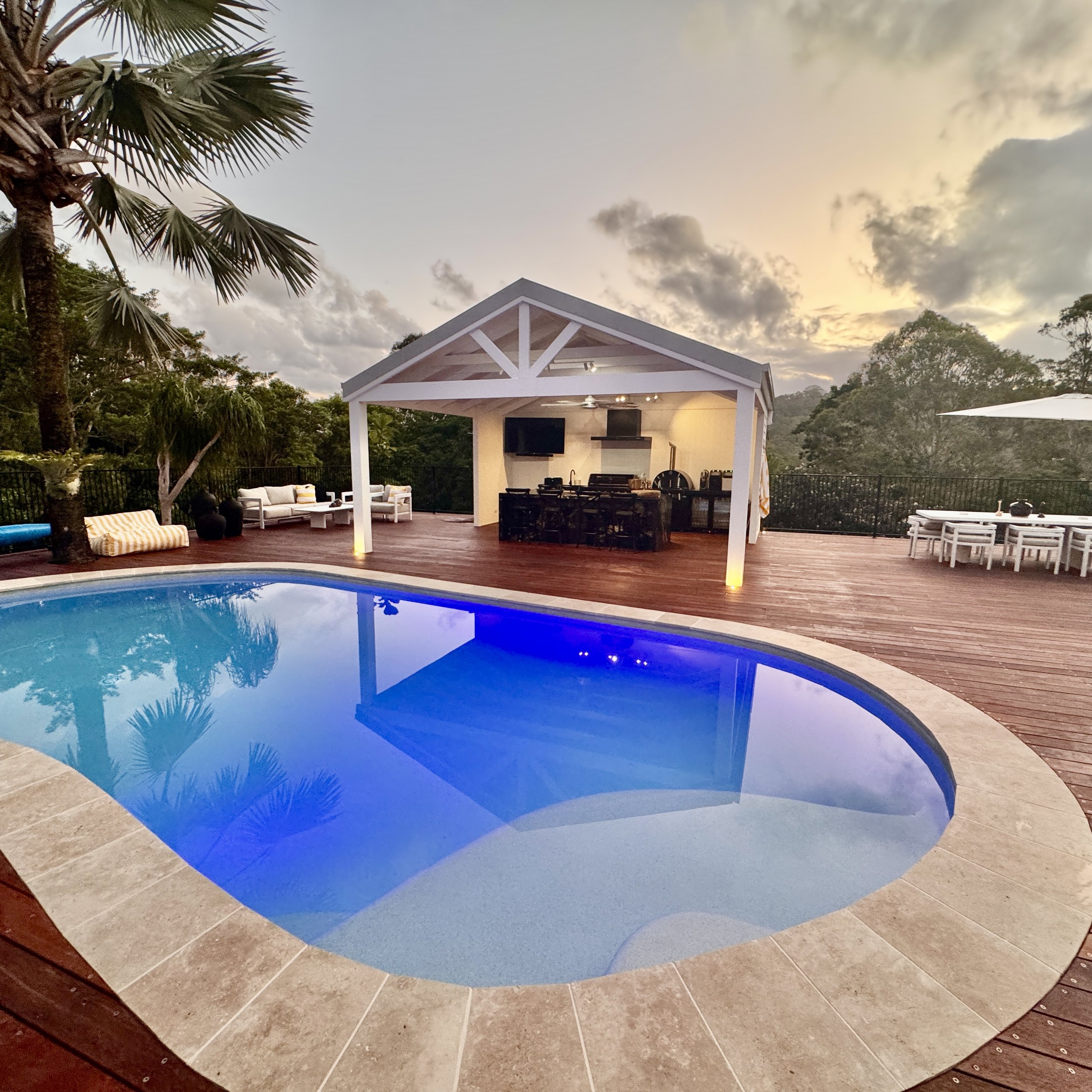 A backyard with a blue illuminated swimming pool, a covered outdoor kitchen and lounge area, and surrounded by trees at sunset.