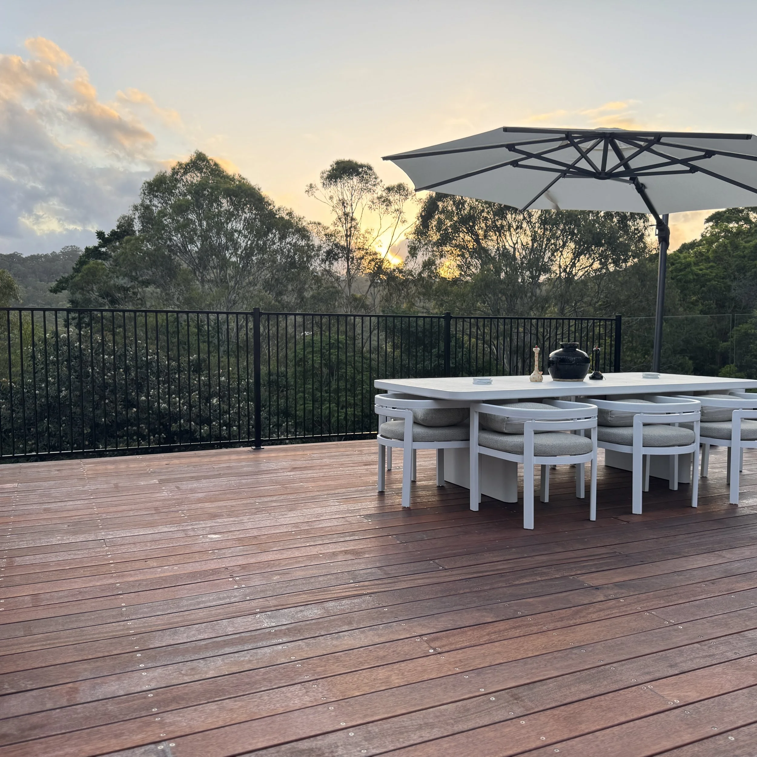 A spacious outdoor deck with wooden planks, black metal railing, a large white umbrella, a white dining table and chairs, and a scenic view of trees at sunset.