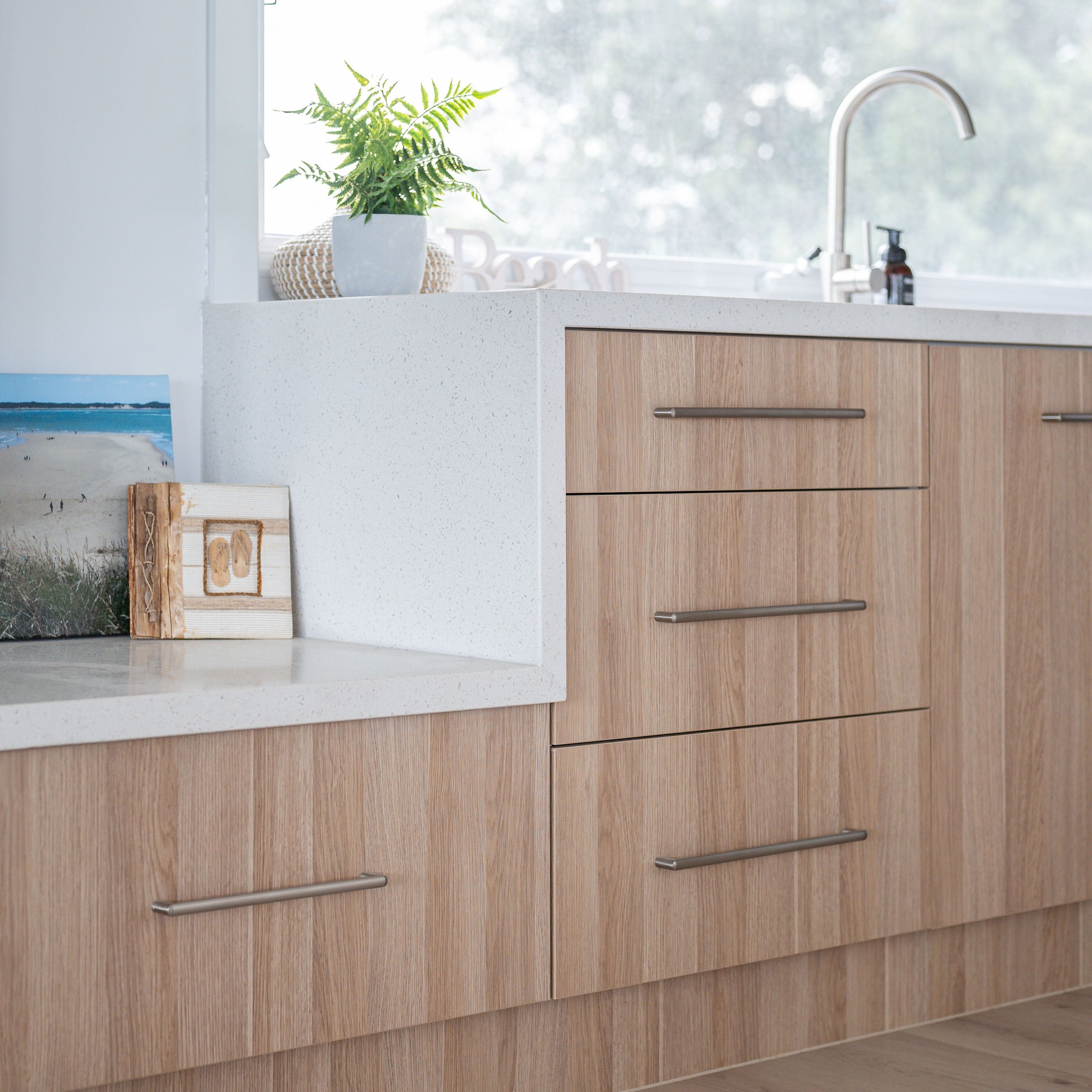 A modern kitchen with wooden cabinets, a white countertop, a plant in a white pot, a painting, and a window in the background.