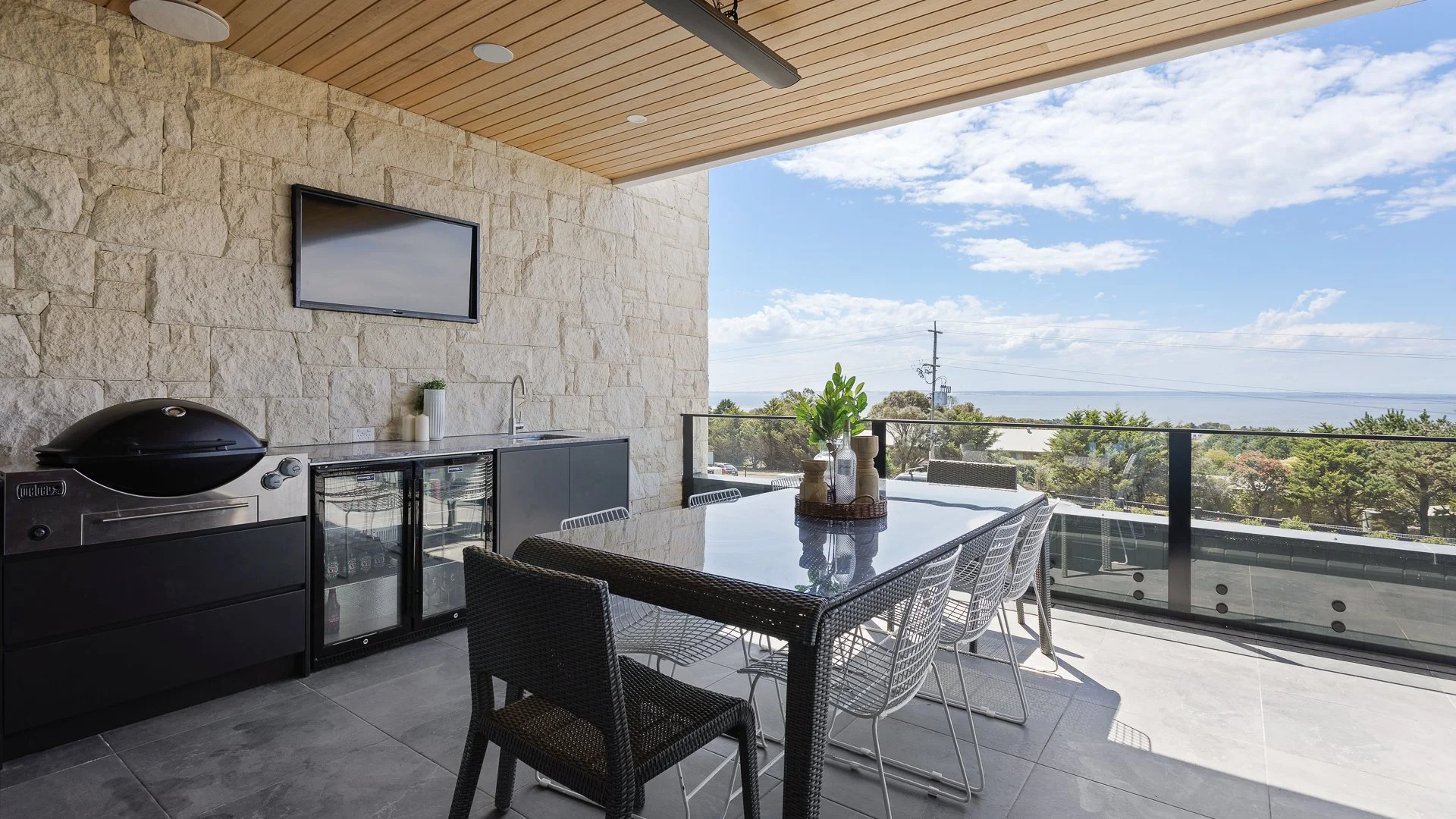 Outdoor balcony with a dining table and chairs, a barbecue grill, a mini fridge, a large TV on a stone wall, and scenic view of trees, power lines, and sky.