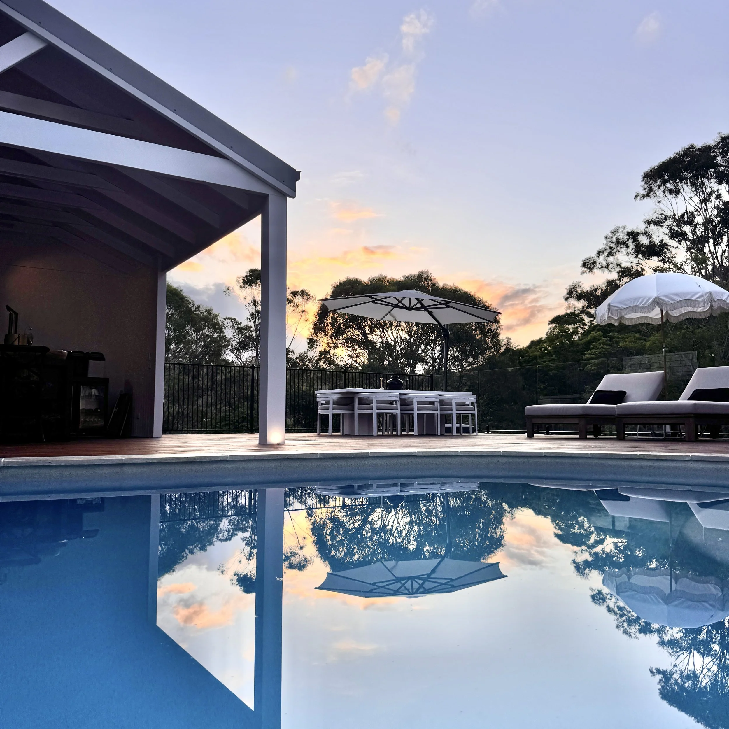 An outdoor swimming pool area at sunset with lounge chairs, umbrellas, a dining table with chairs, and trees in the background, reflected in the pool water.
