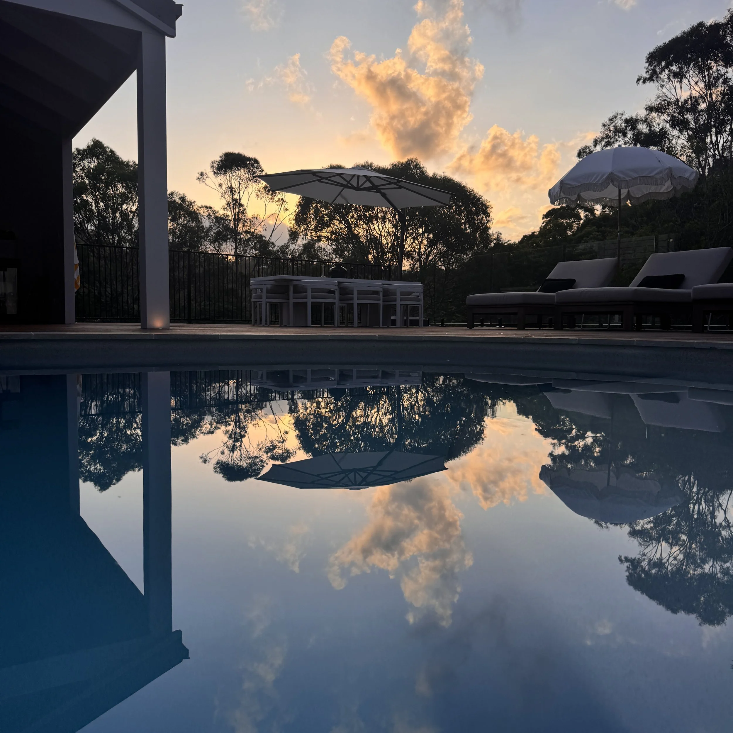 A backyard pool at sunset with outdoor furniture and umbrellas, reflecting the sky and trees in the water.