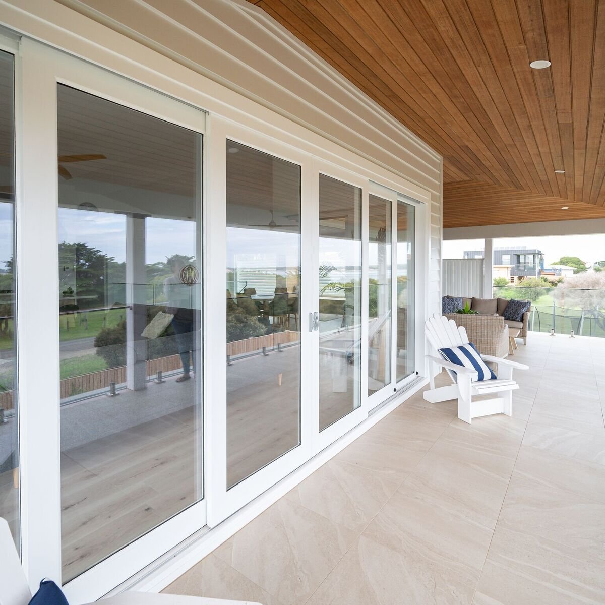 Empty balcony with glass sliding doors, wicker sofa with cushions, white Adirondack chair with striped pillow, and a view of greenery and neighboring houses in the background.