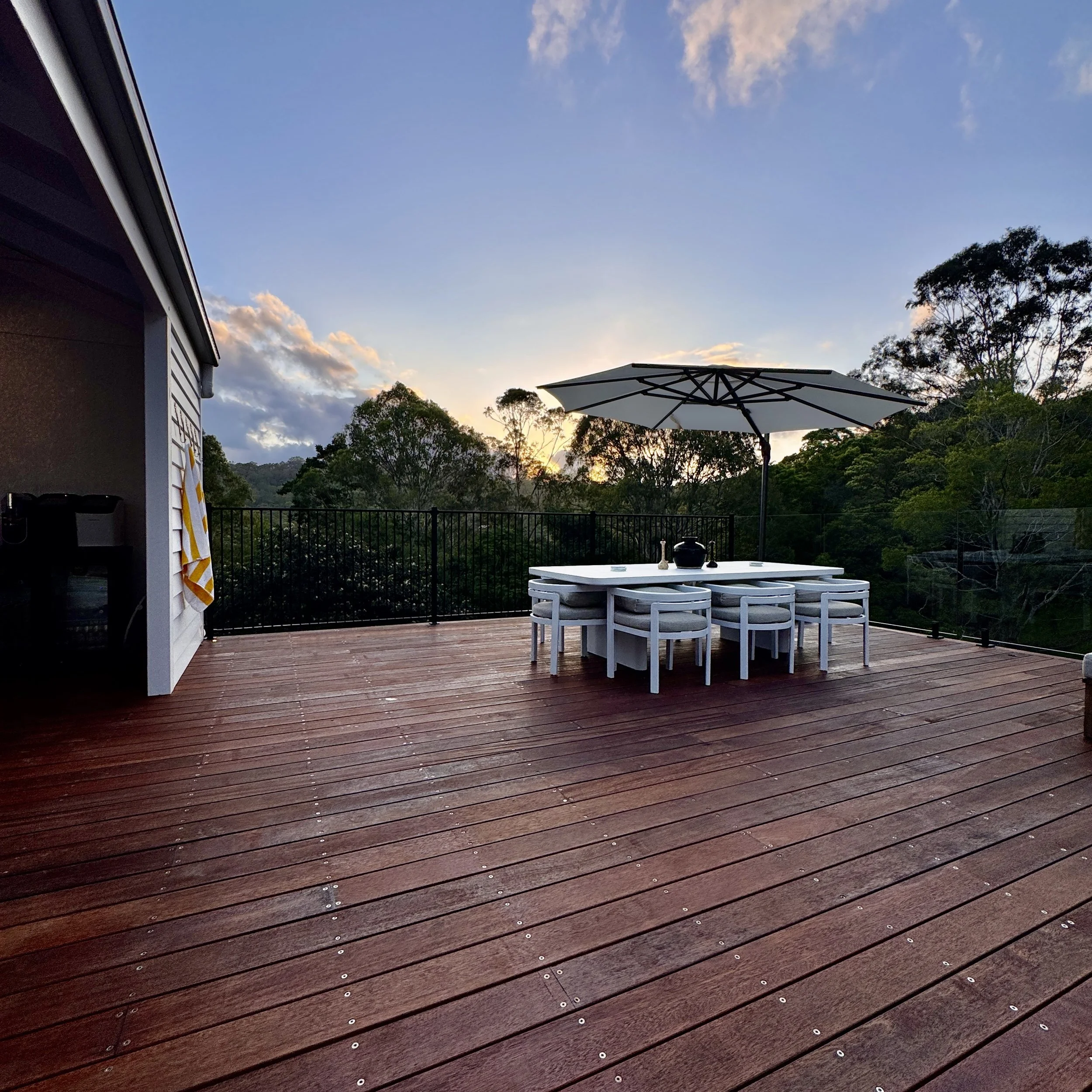 An outdoor deck with wooden flooring, a white dining table with six chairs, and a large umbrella, overlooking a green landscape at sunset.