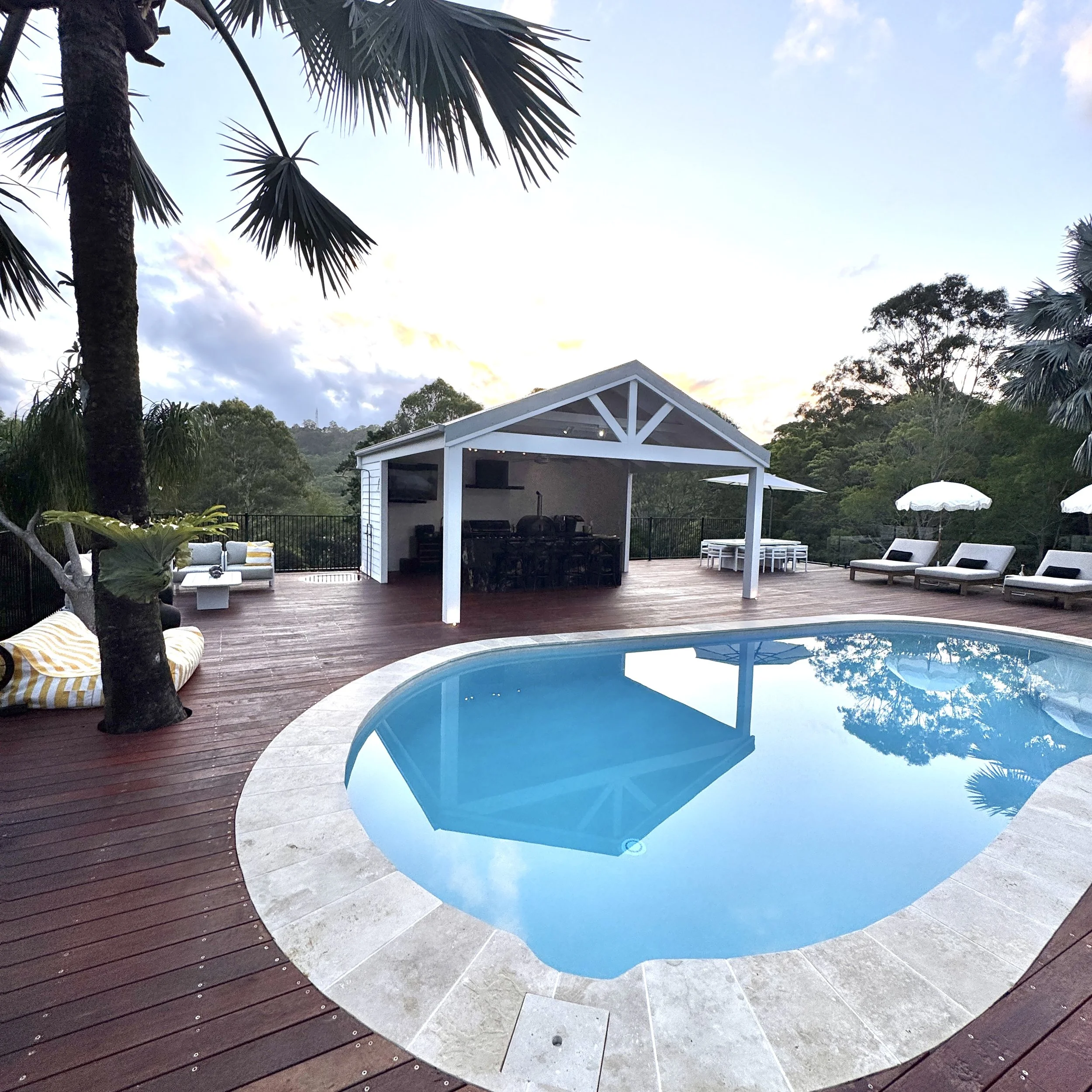 A backyard swimming pool with a wooden deck, surrounded by lounge chairs with umbrellas, trees, and a small white pavilion with a bar area in the background, set against a scenic landscape with hills and a sky during sunset.