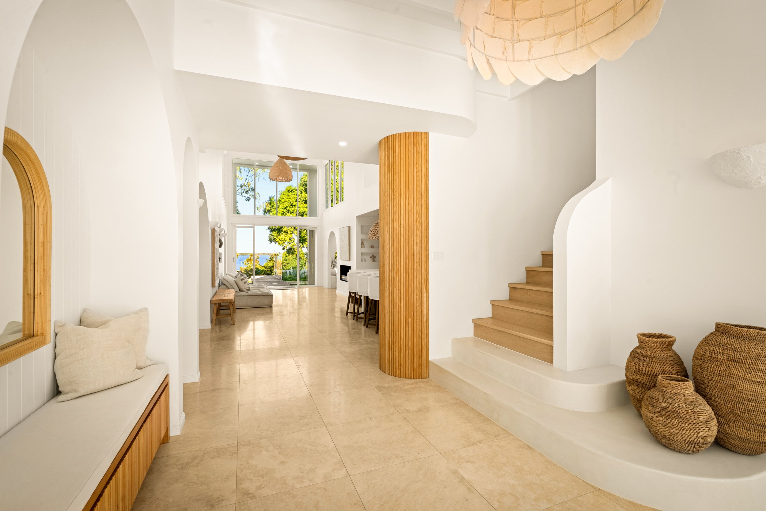 Interior of a modern, airy home featuring white walls, wooden accents, a staircase, woven baskets, a sitting area with a view of trees and the ocean.