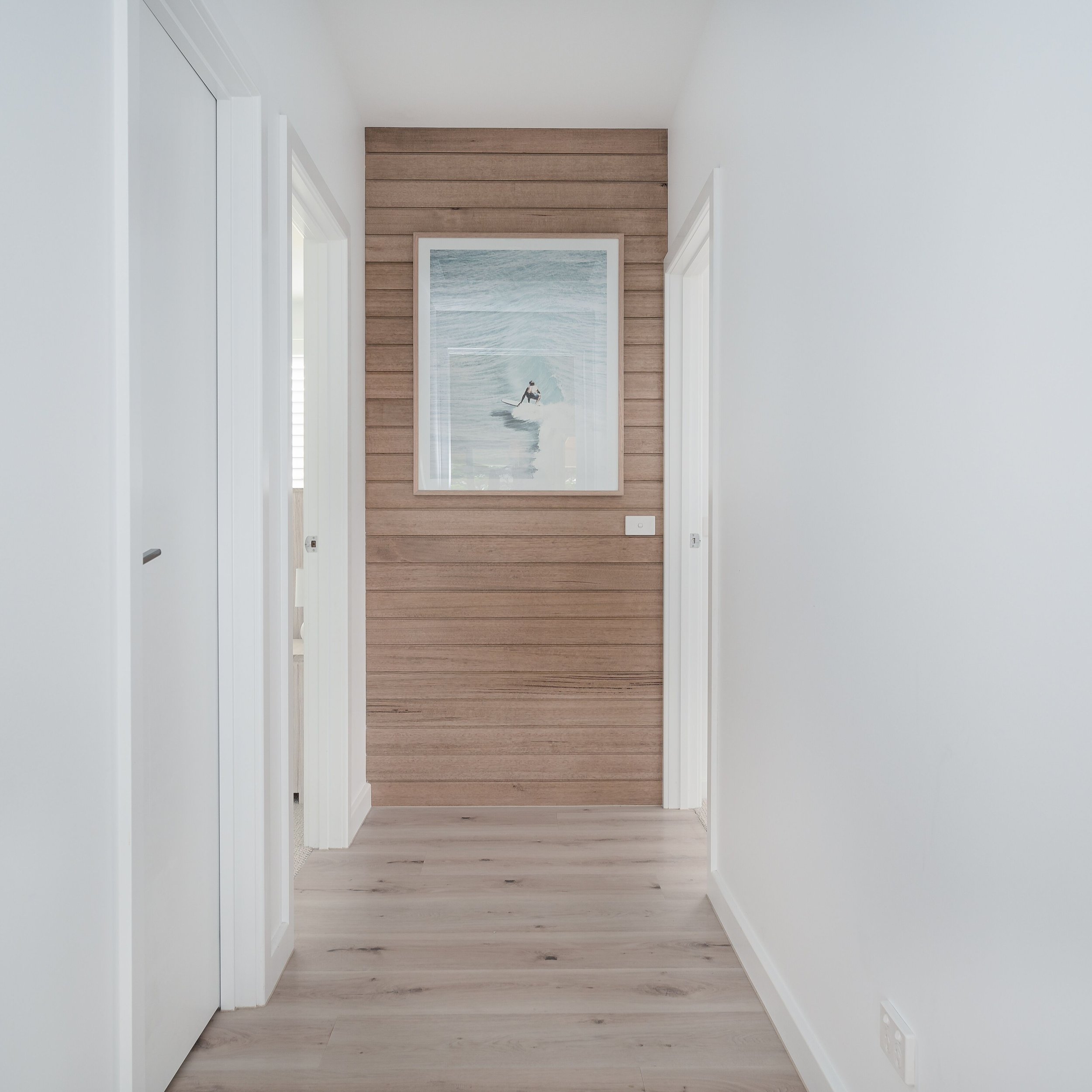Minimalist hallway with white walls, light wood flooring, and a wood-paneled accent wall with a framed picture of a surfer in the ocean.