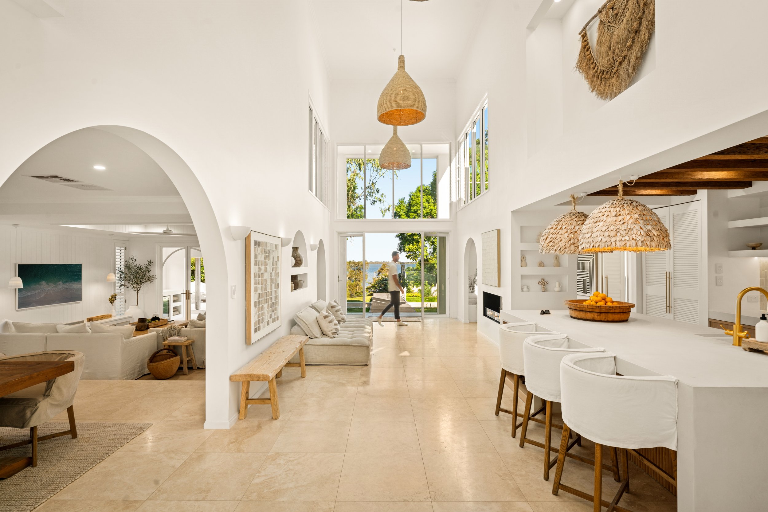Bright, airy living room and kitchen with white walls, large windows, hanging pendant lights, beige tiled floor, and minimalist decor.
