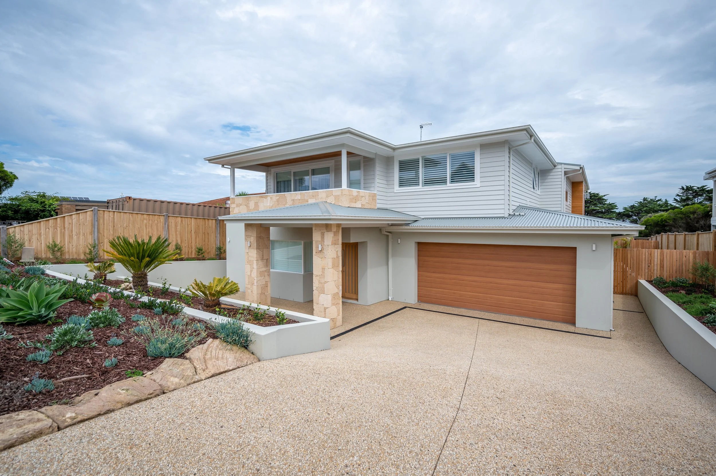 Modern two-story house with white siding, stone accents, and a wooden garage door, surrounded by a landscaped garden and a paved driveway under a partly cloudy sky.