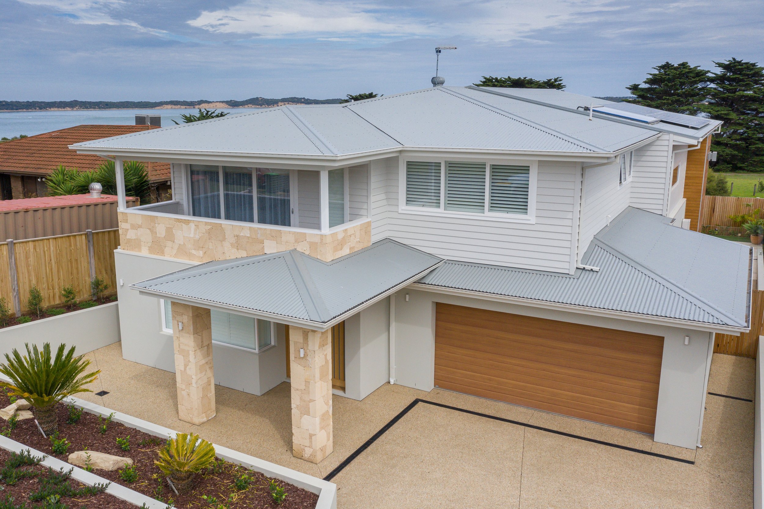 Modern two-story house with white siding, stone accents, and a wooden garage door, with a landscaped front yard and a view of water in the background.