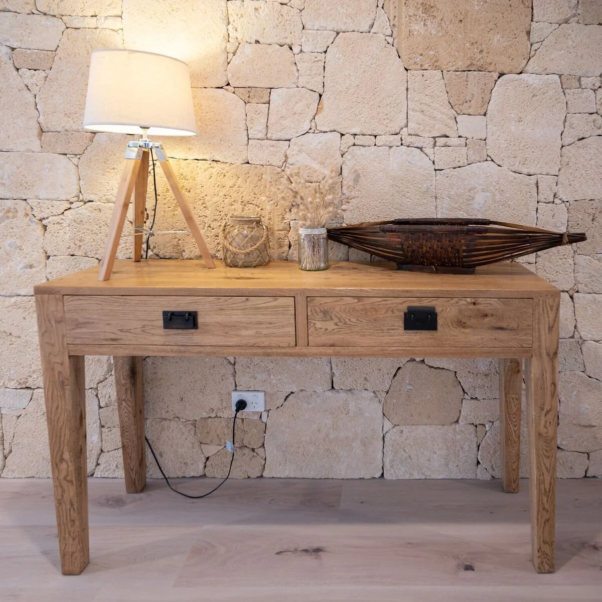 A wooden console table against a stone wall, topped with a white table lamp, decorative jars, a dried plant arrangement, and a woven boat-shaped decor piece. An electrical outlet with a plugged-in cord is visible beneath the table.