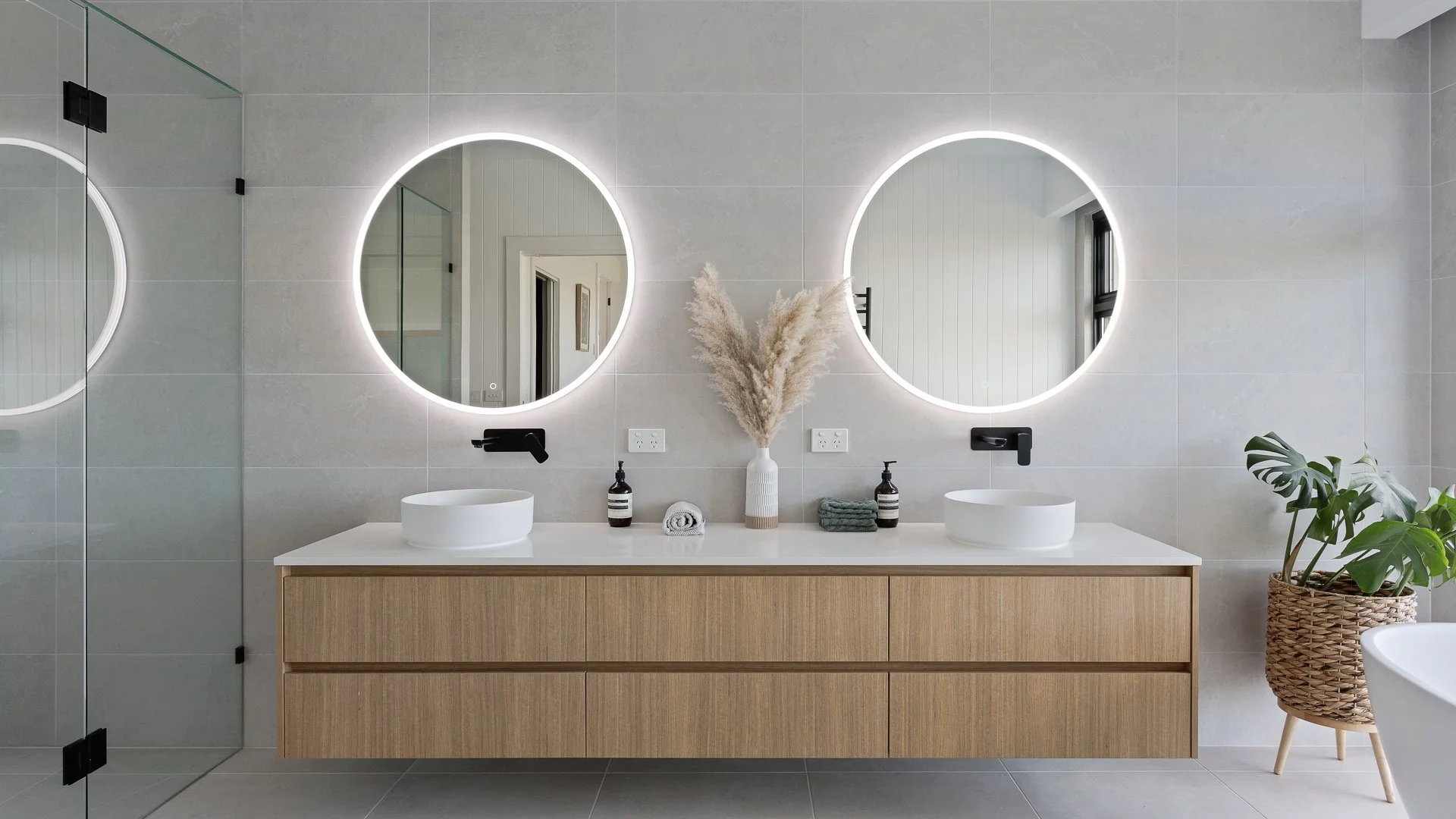 Modern bathroom with a double vanity, two round illuminated mirrors, and neutral-colored decor, including a vase with pampas grass and a potted plant.