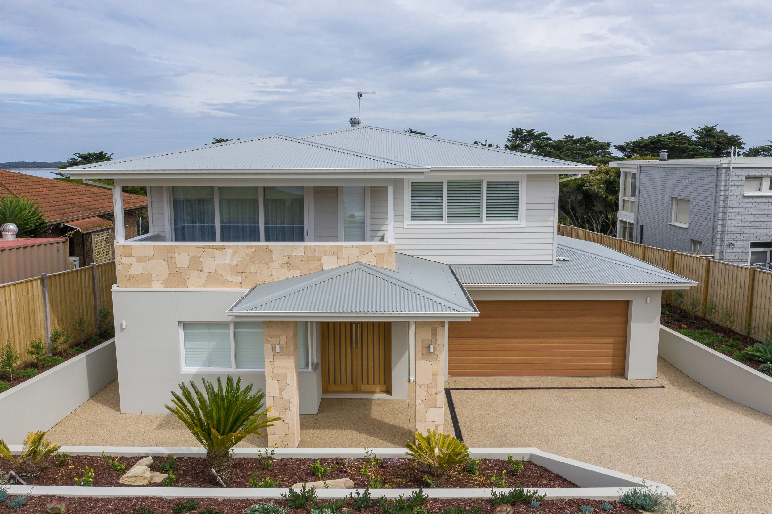Modern two-story house with stone and white siding exterior, gray metal roof, wooden garage door, small front yard with plants, and fenced backyard.