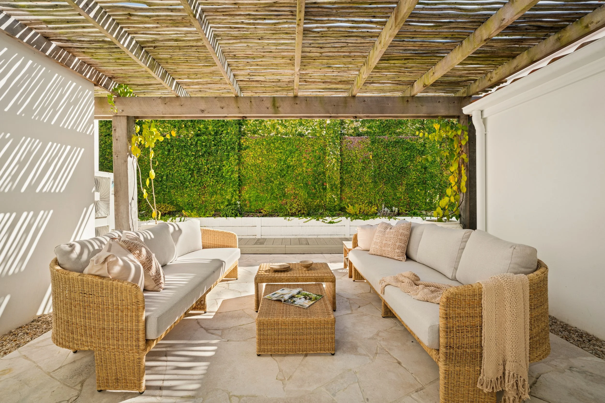 Outdoor patio with two rattan sofas with white cushions, a wicker coffee table with magazines and bowls, under a wooden pergola with a lattice roof, and a green hedge wall in the background.
