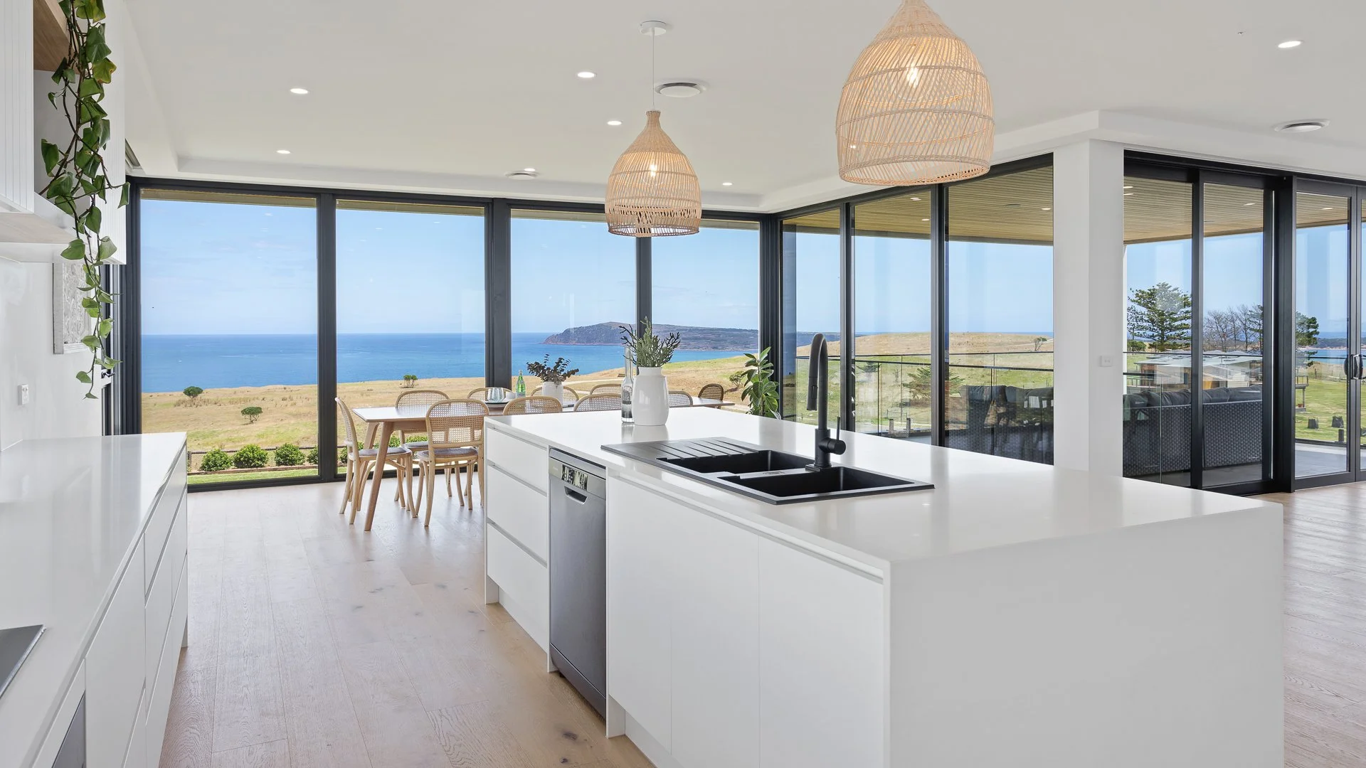 Modern kitchen with white cabinets, black sink, large windows with ocean view, dining table outside, decorative plants, wicker pendant lights.