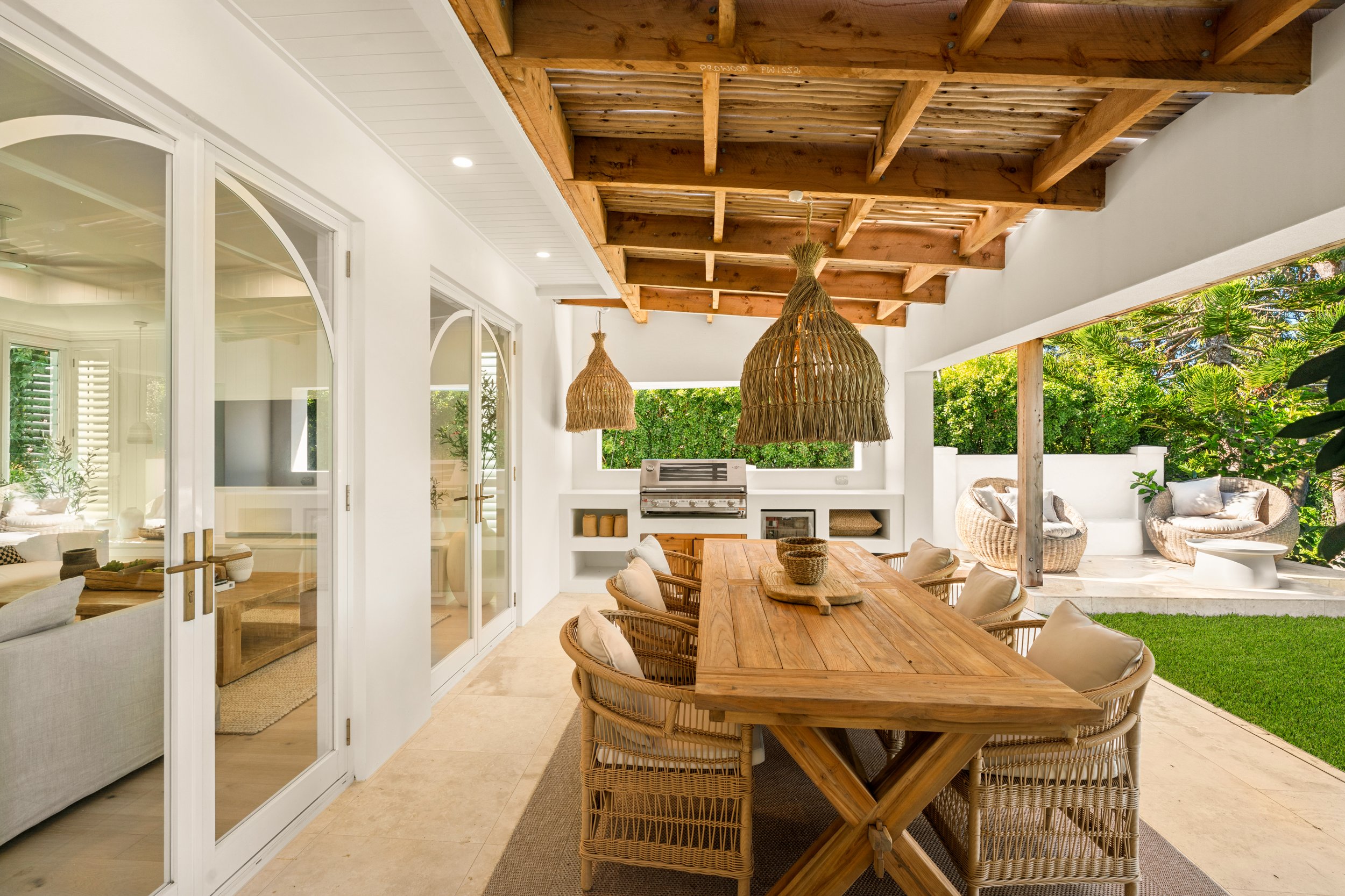 Outdoor patio with wooden dining table surrounded by wicker chairs, hanging woven pendant lights, built-in barbecue, and seating area with cushioned wicker chairs, overlooking a green garden.