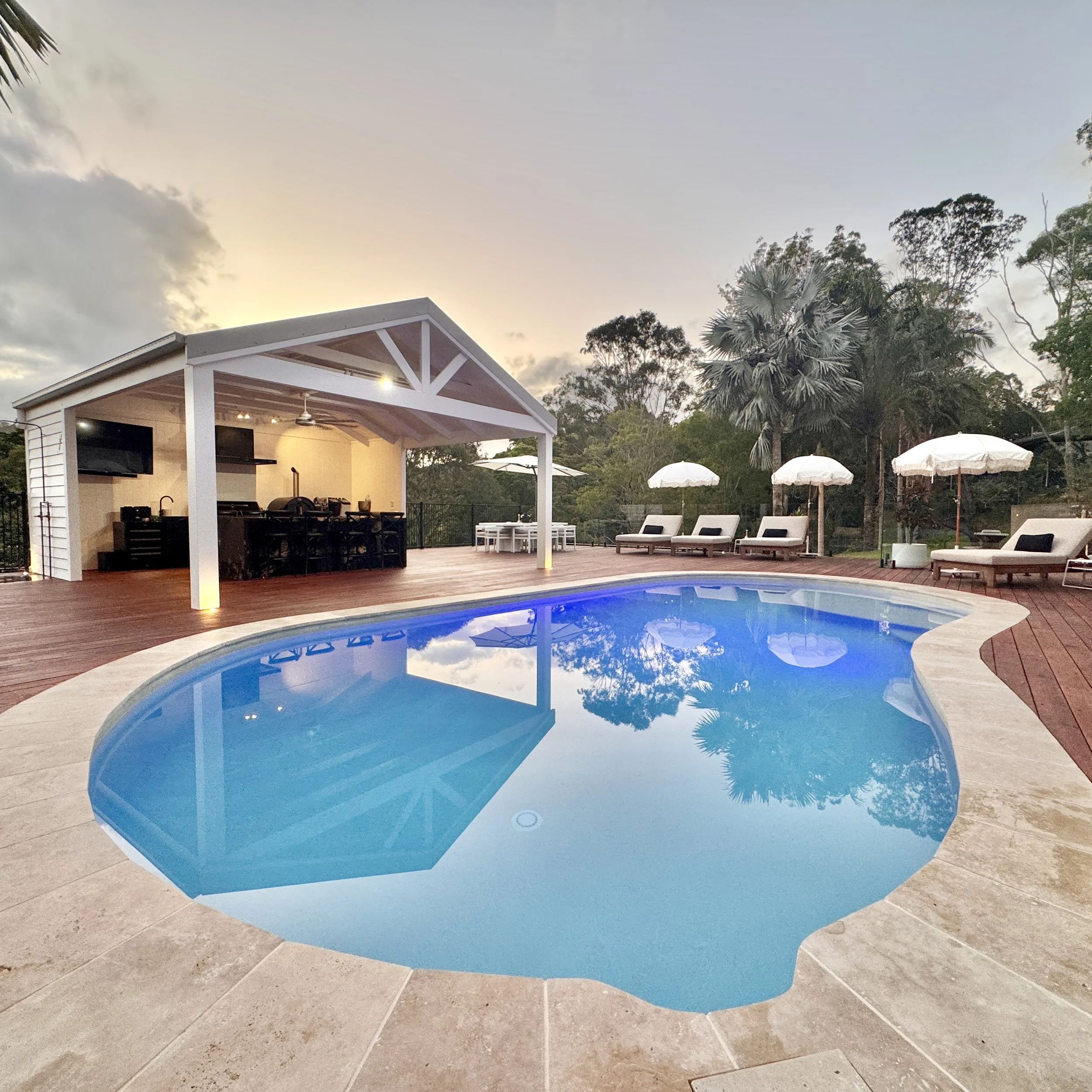 A backyard with a swimming pool, white poolside loungers with black cushions, white umbrellas, and a covered outdoor kitchen and dining area surrounded by tall trees.