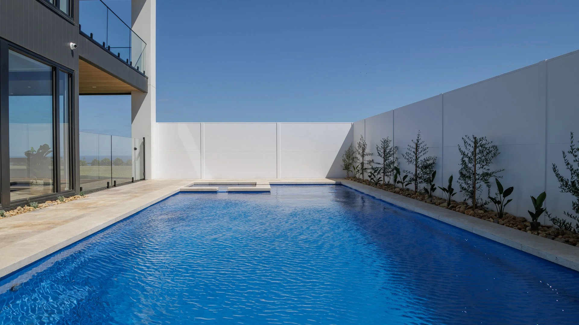 Modern house with large glass doors and a private swimming pool surrounded by a white wall, with a clear blue sky overhead.