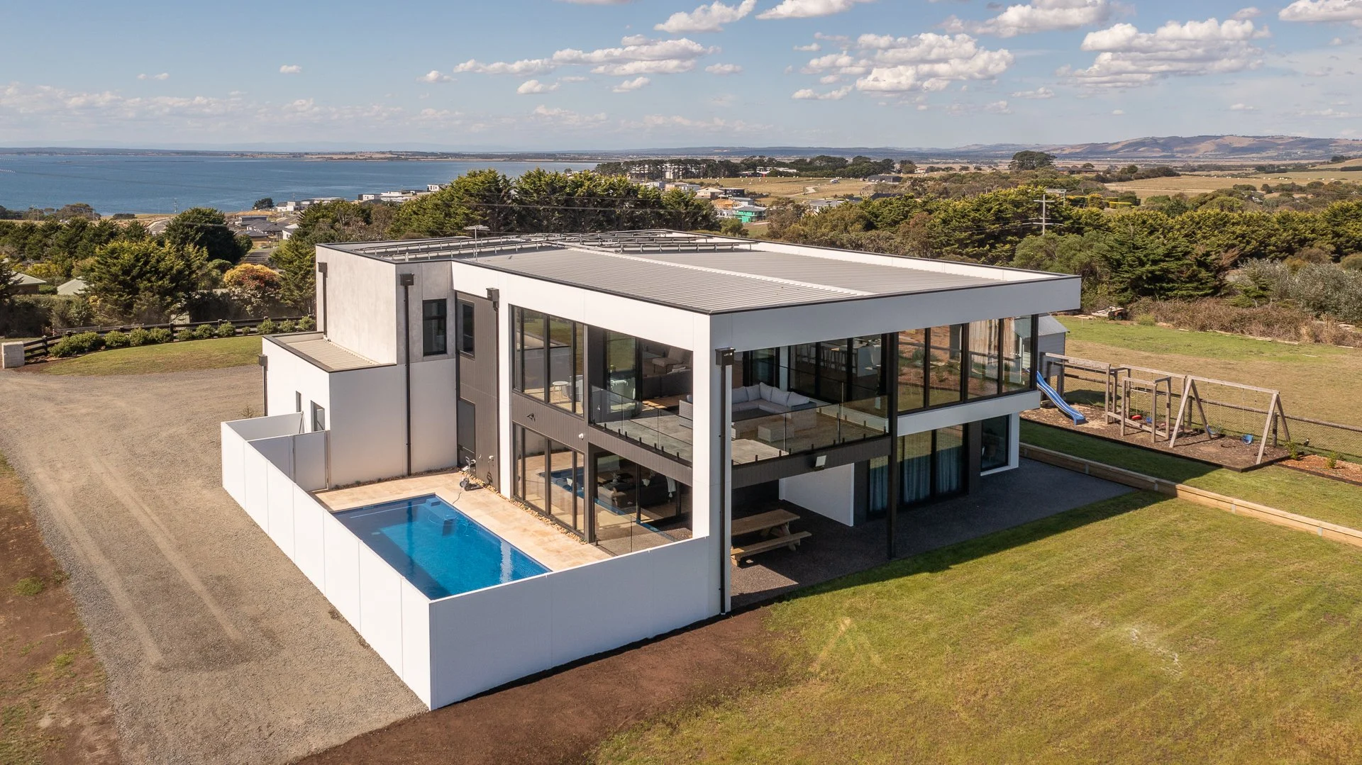 Modern two-story house with large glass windows, a backyard swimming pool, a backyard swing set, and a surrounding lawn with trees overlooking a body of water in the distance under a partly cloudy sky.