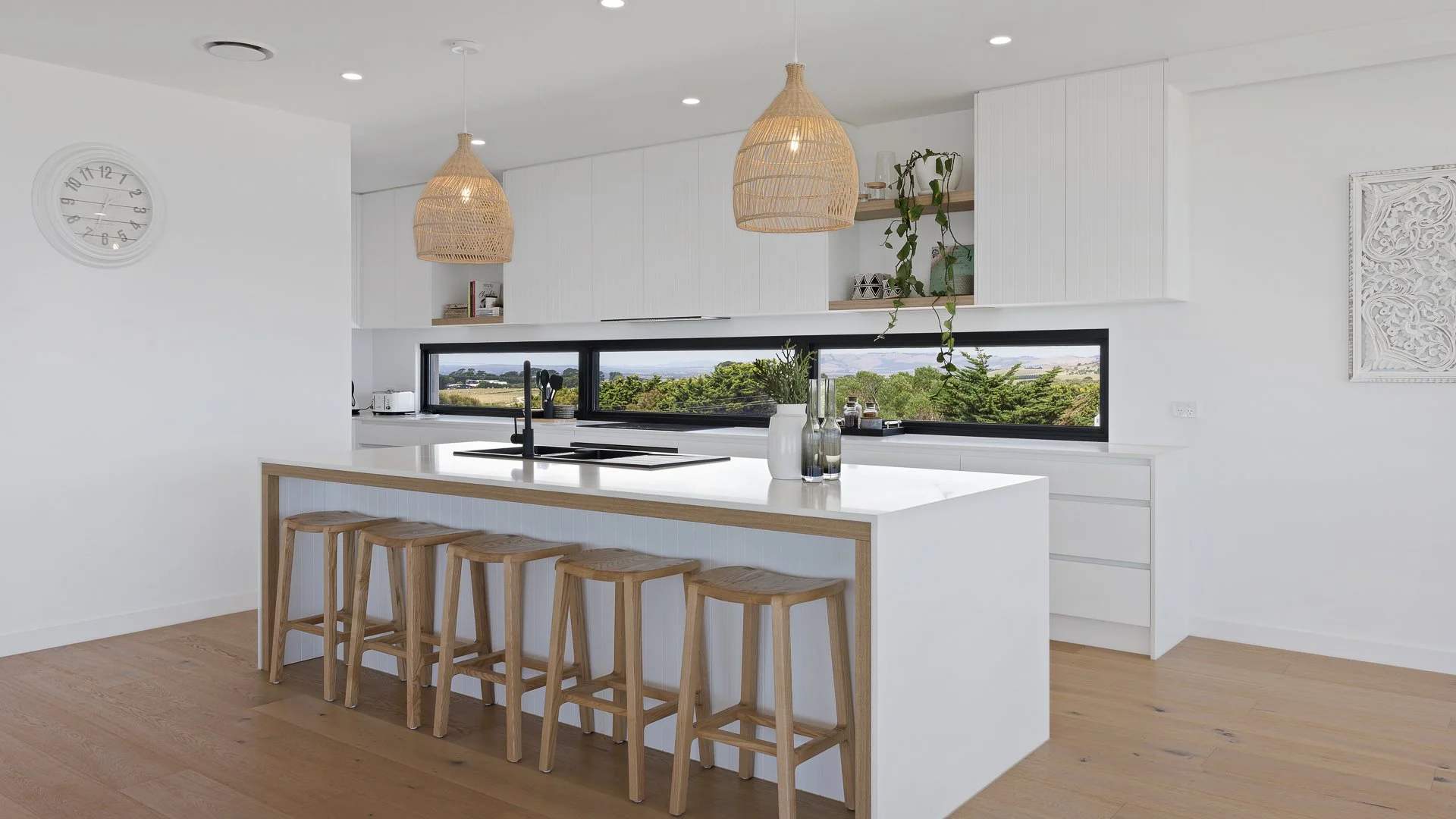 Bright modern kitchen with white cabinets, a white island with wooden bar stools, a black window, hanging wicker pendant lights, and a wall clock.