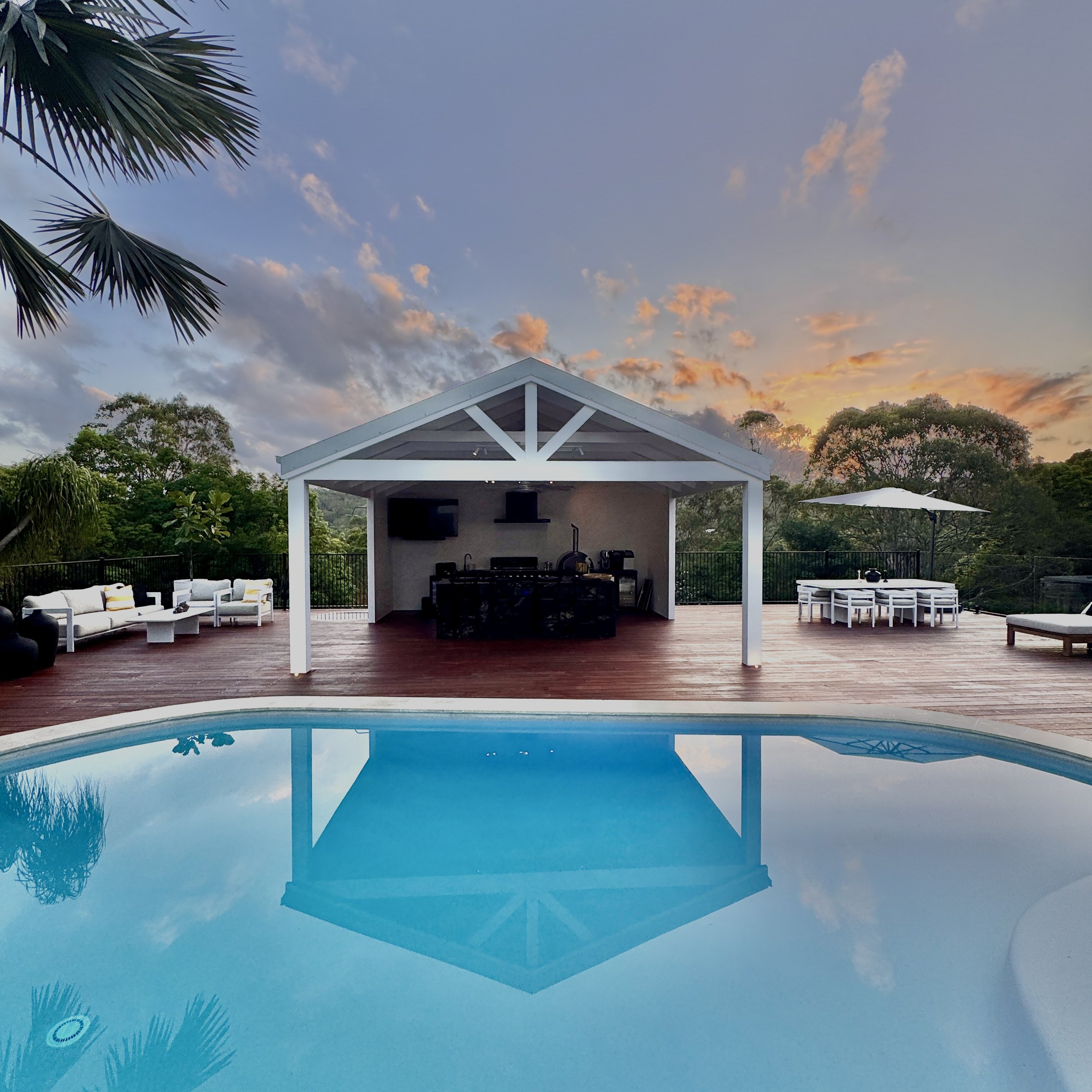 A backyard pool area at sunset with a pool in the foreground, a covered outdoor kitchen and seating area with a TV, and outdoor dining with an umbrella, surrounded by trees and a wooden deck.