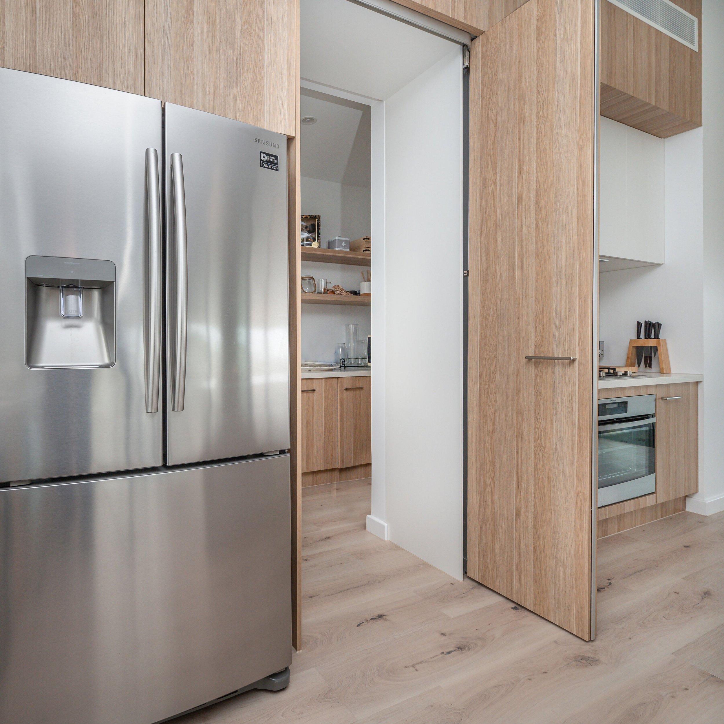 Modern kitchen with stainless steel refrigerator, wooden cabinetry, and built-in oven. Partially open sliding door reveals a small pantry or storage area with shelves.