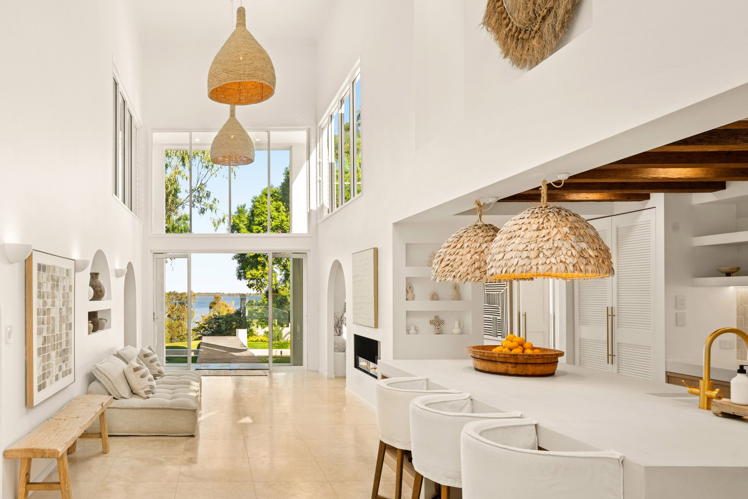 Bright, airy living space with large windows, white walls, beige floor tiles, a bench with cushions, built-in shelves with decor, a fireplace, and a kitchen with white counter and barstools, featuring natural woven pendant lights and a view of outdoo