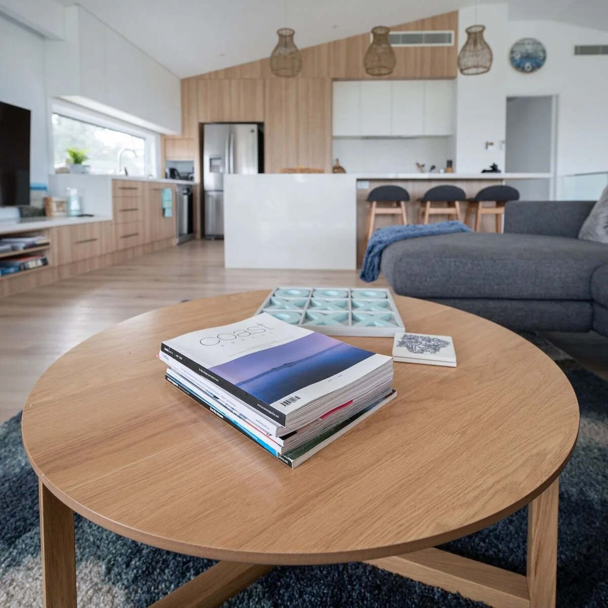 A living room with a wooden coffee table, magazines and books on top, a grey sofa with a blue throw blanket, a kitchen in the background with barstools, pendant lights, a stainless steel refrigerator, and a window with a small potted plant.