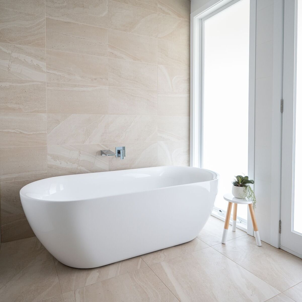 Modern bathroom with beige tiled wall and floor, white freestanding bathtub, small white stool with potted plant, and large glass door letting in natural light.