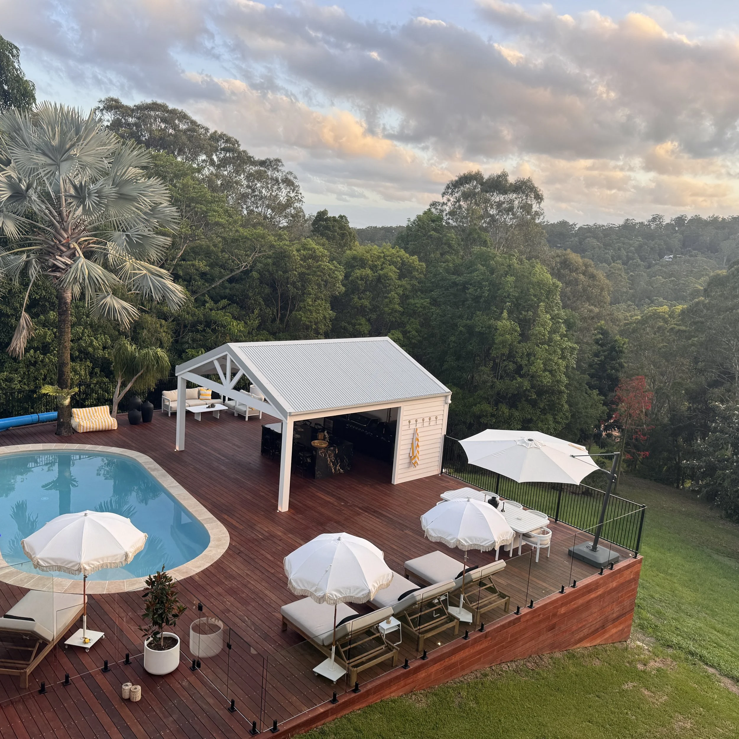 A backyard pool area with a swimming pool, white umbrellas, lounge chairs, a covered outdoor bar area, and lush green trees in the background during late afternoon or early evening.