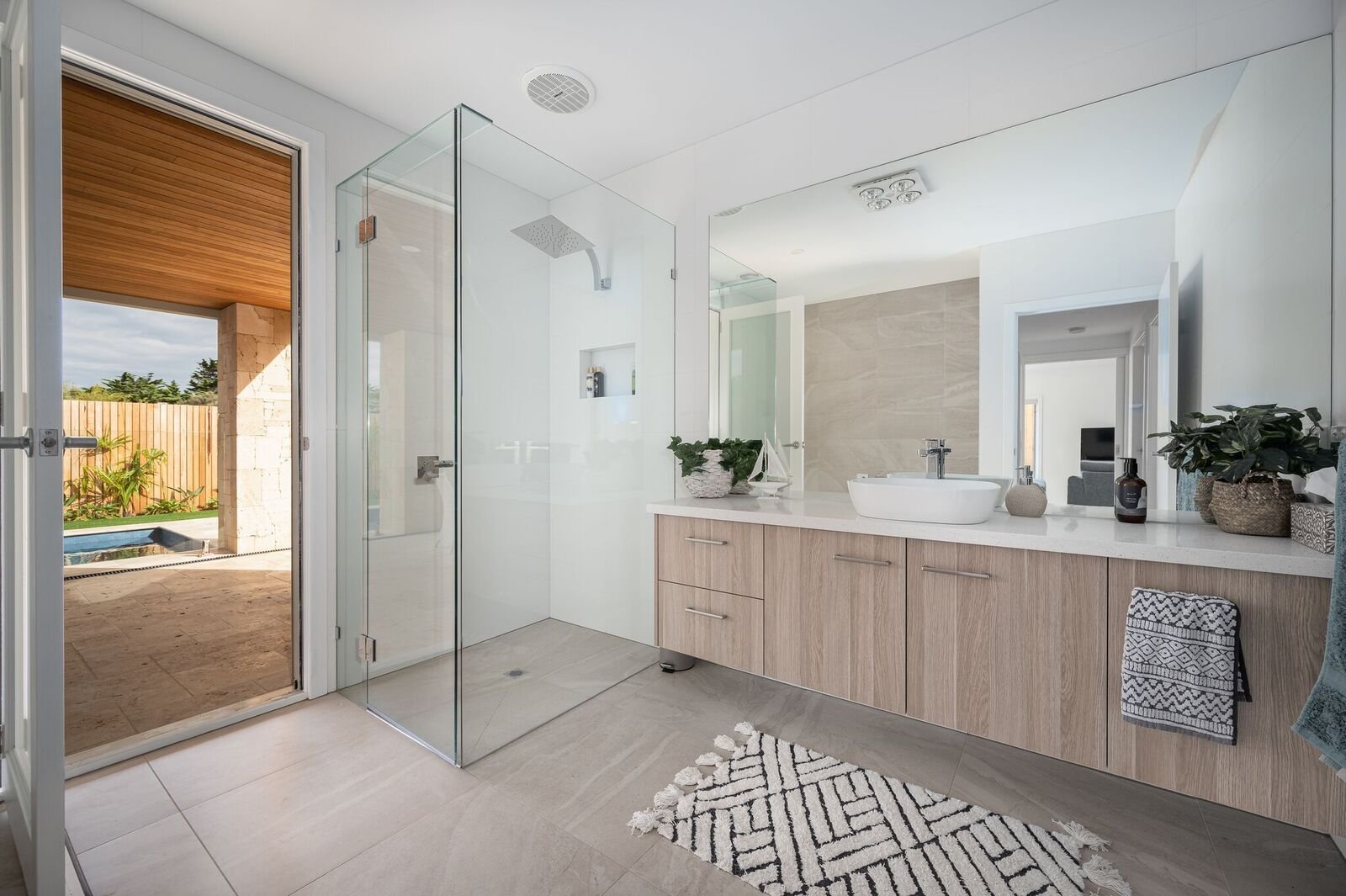 Modern bathroom with glass shower enclosure, wooden vanity with white countertop, large mirror, and door opening to outdoor patio with swimming pool.
