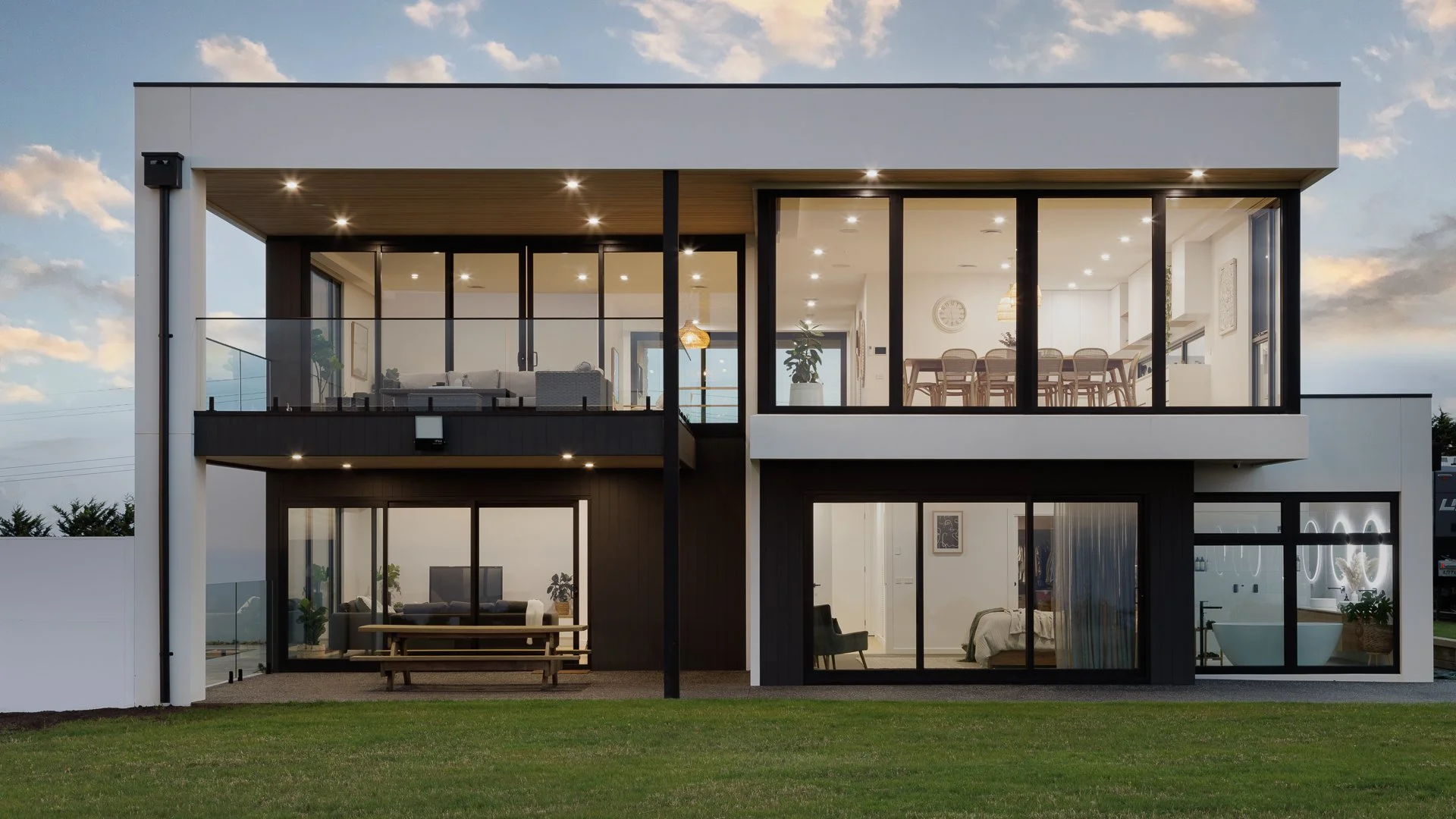 Modern two-story house with large glass windows and balconies, illuminated from within, with a green lawn in the foreground and a cloudy sky in the background.