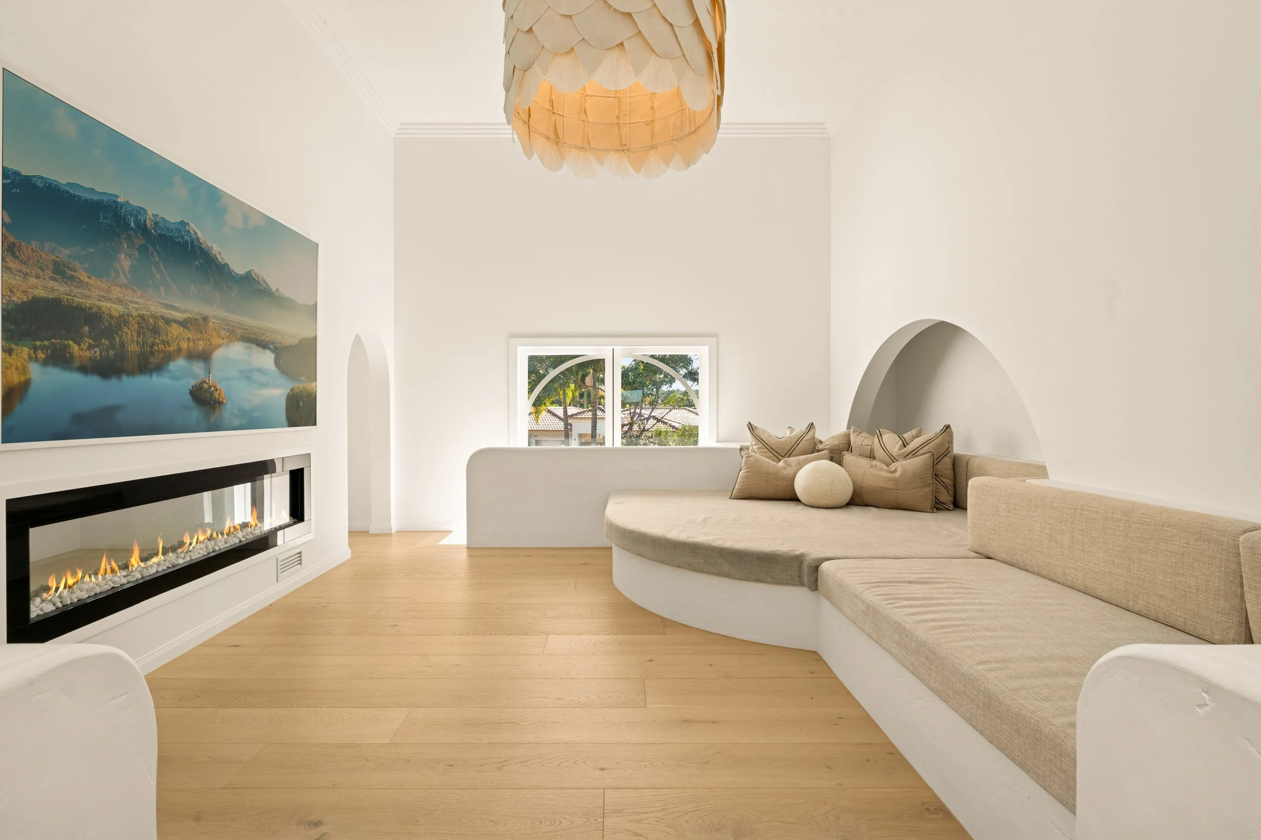 Minimalist living room with white walls, a built-in fireplace, a large landscape photograph on the wall, a curved beige and tan couch with pillows, and a window showing greenery outside. A hanging light fixture is on the ceiling.