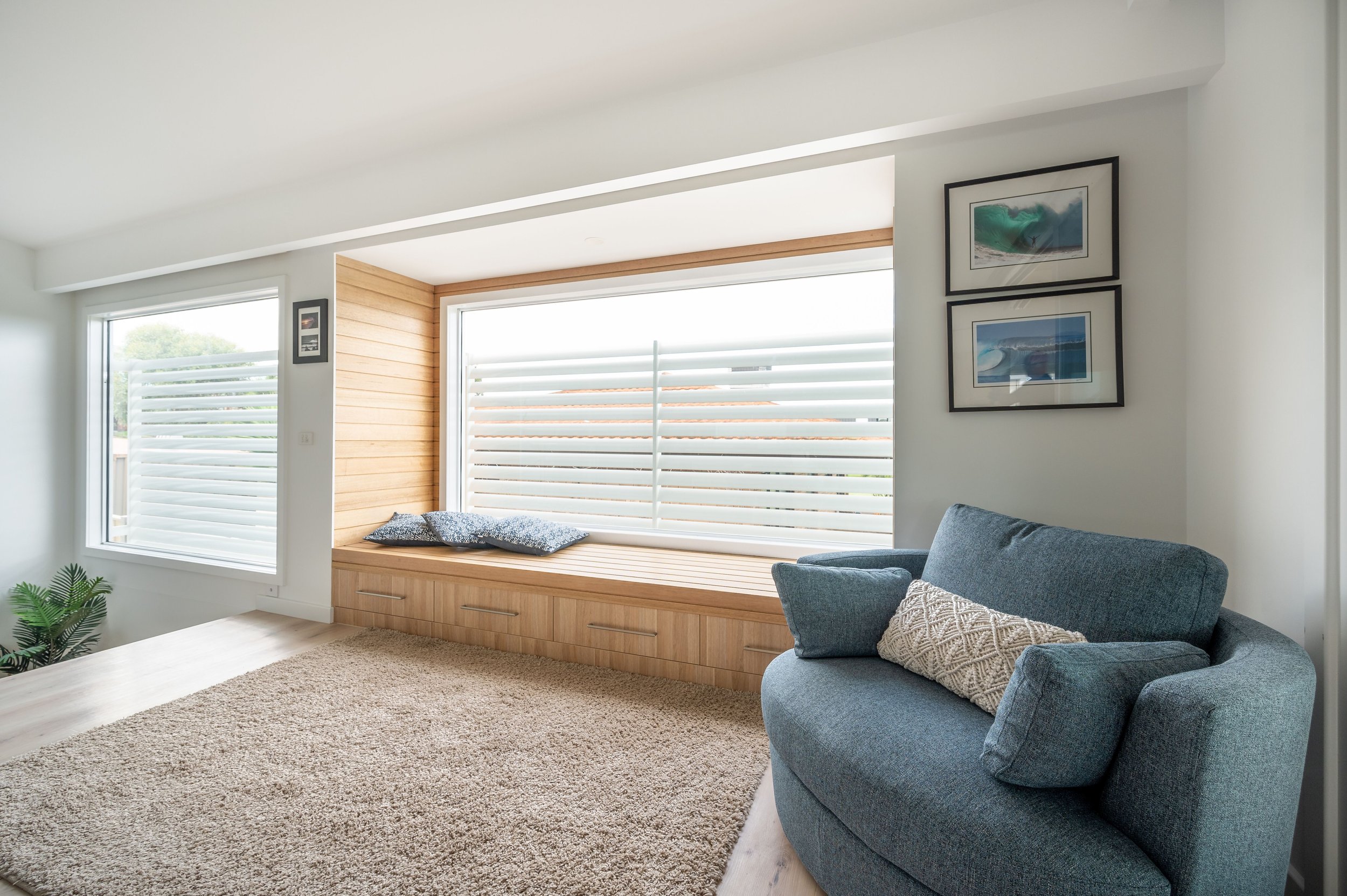 Living room with a large window seat with pillows, a blue armchair with a patterned cushion, framed pictures on the wall, a window with sliding shutters, a beige carpet, and a small plant in the corner.