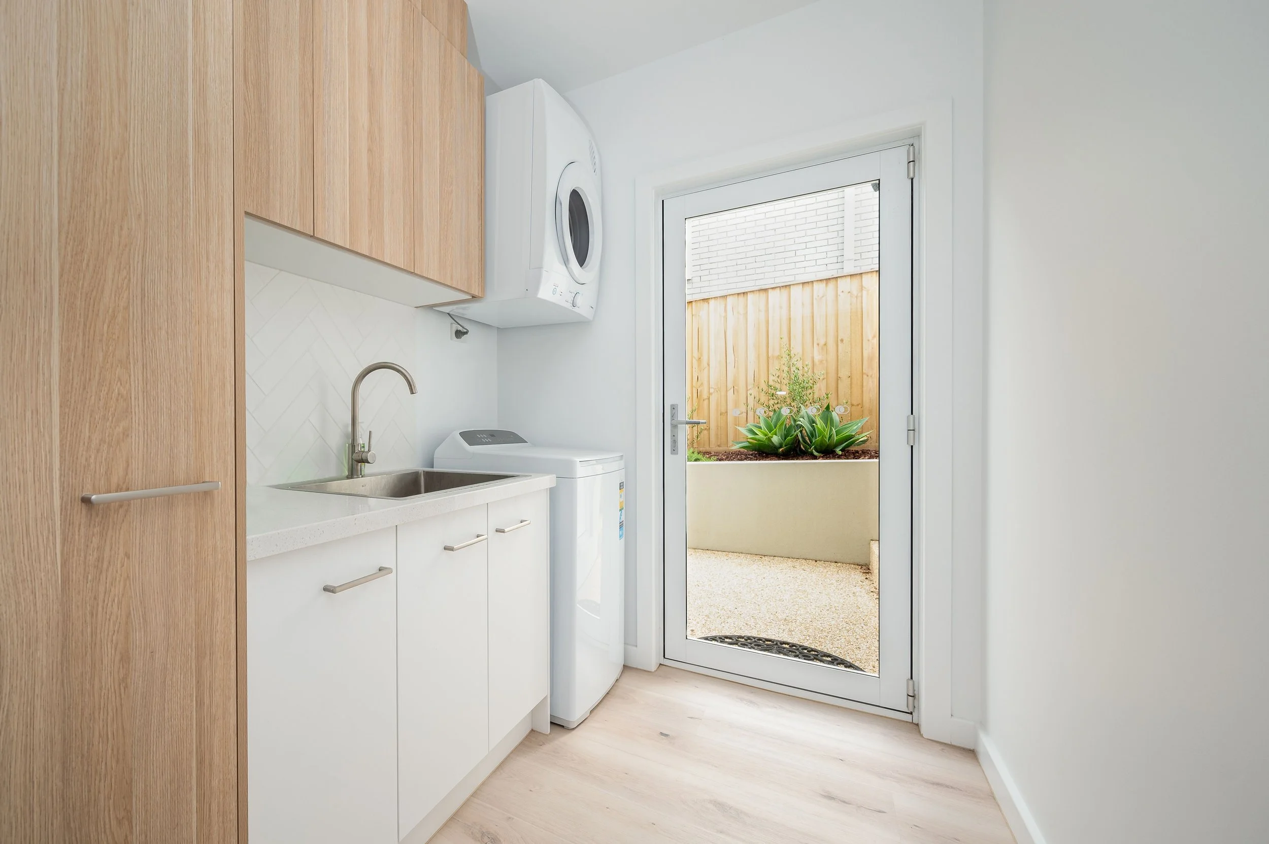 Laundry room with white cabinets, a washing machine, a dryer, a sink, and a door leading outside to a small patio with plants.