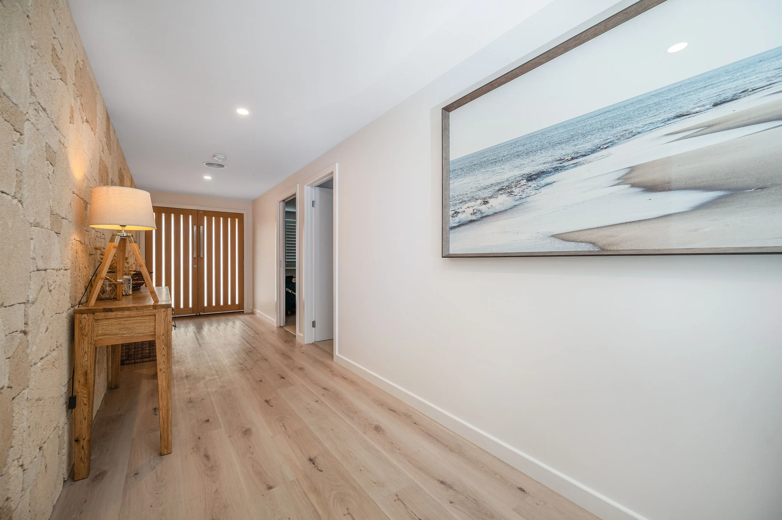 A hallway with light hardwood flooring, white walls, and a brick accent wall on the left. There is a wooden console table with a lamp on it near the brick wall. A large framed painting of a beach scene hangs on the right wall. Doors and a brown woode