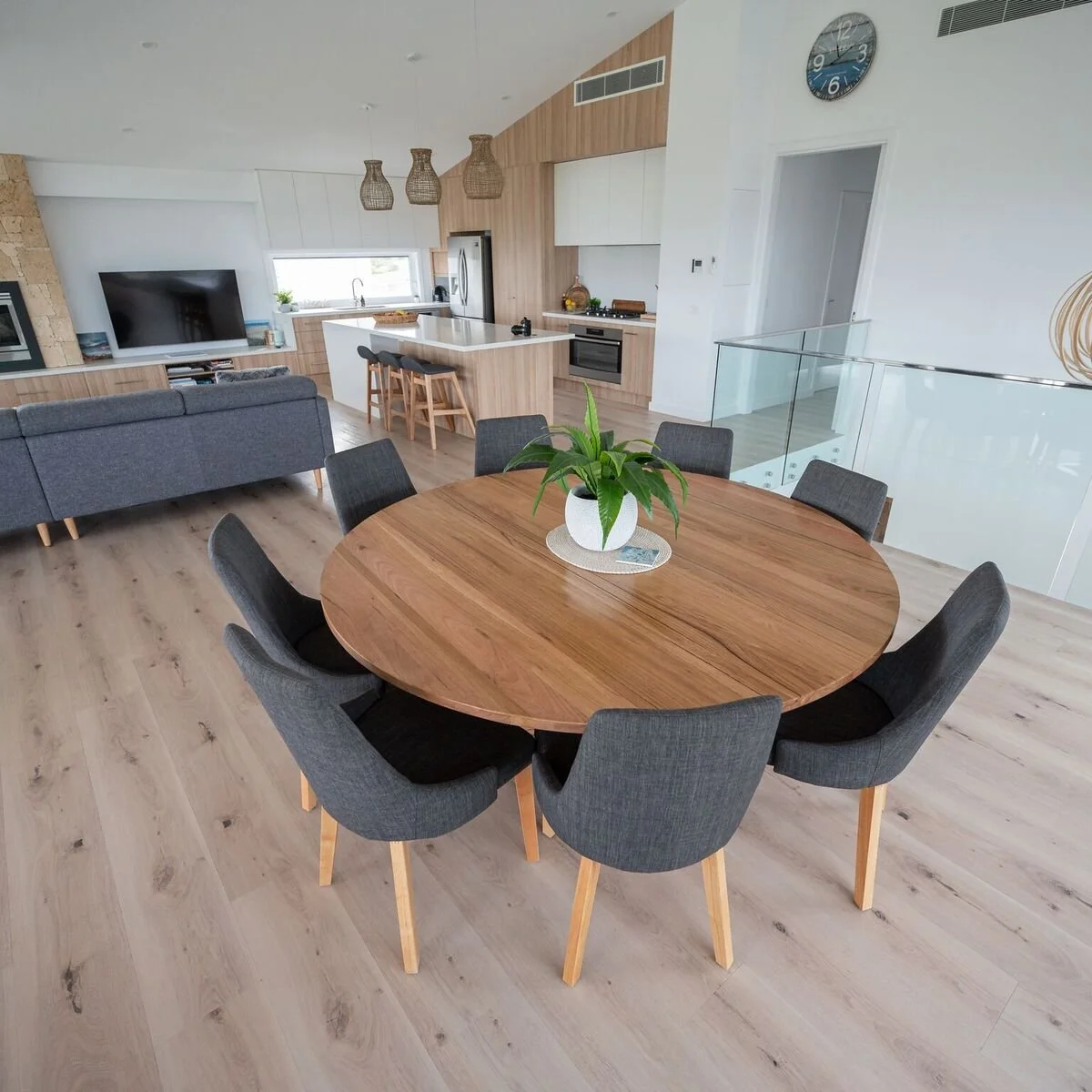A modern open-concept living space with a round wooden dining table surrounded by six dark gray upholstered chairs, a potted plant in the center, and a kitchen area with a breakfast bar, gray sofa, flat-screen TV, and wooden cabinets in the backgroun