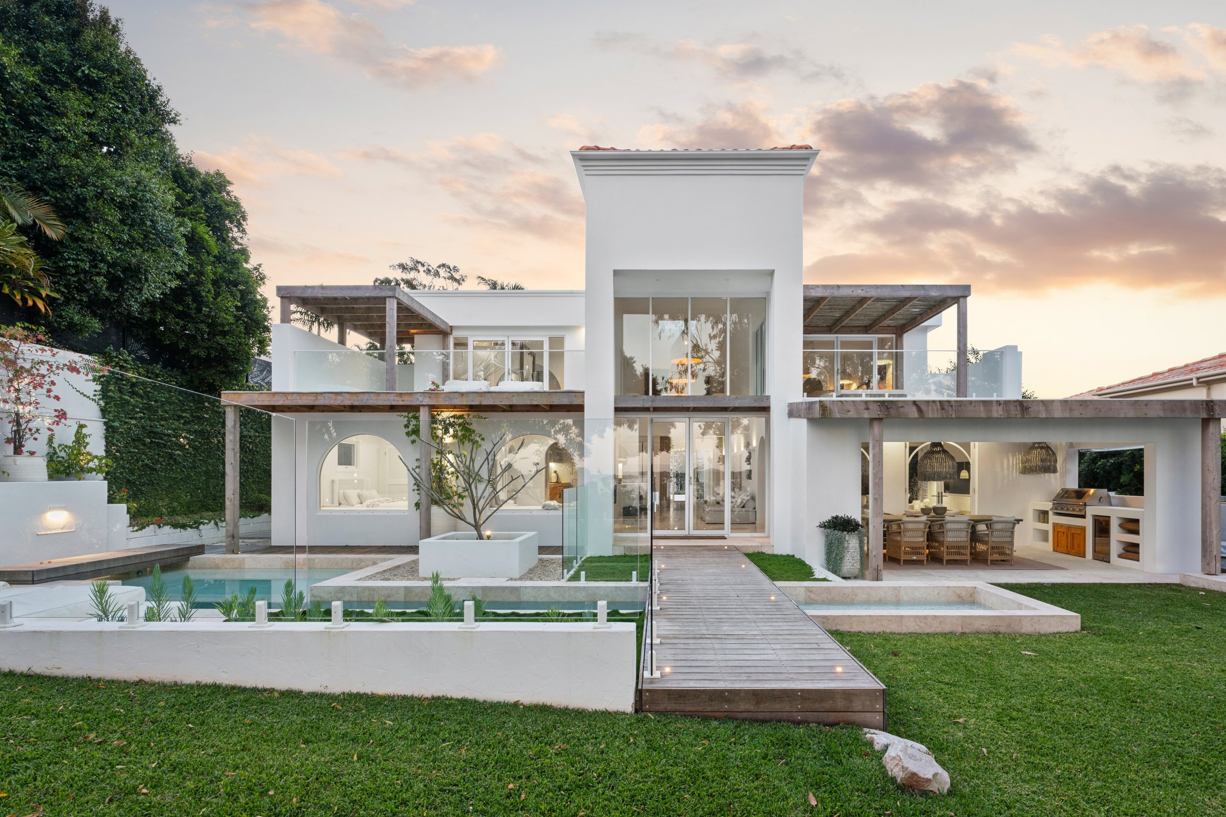 Modern white house with large glass windows and a flat rooftop, with a backyard featuring a small pool, grassy lawn, and outdoor dining area, during sunset.