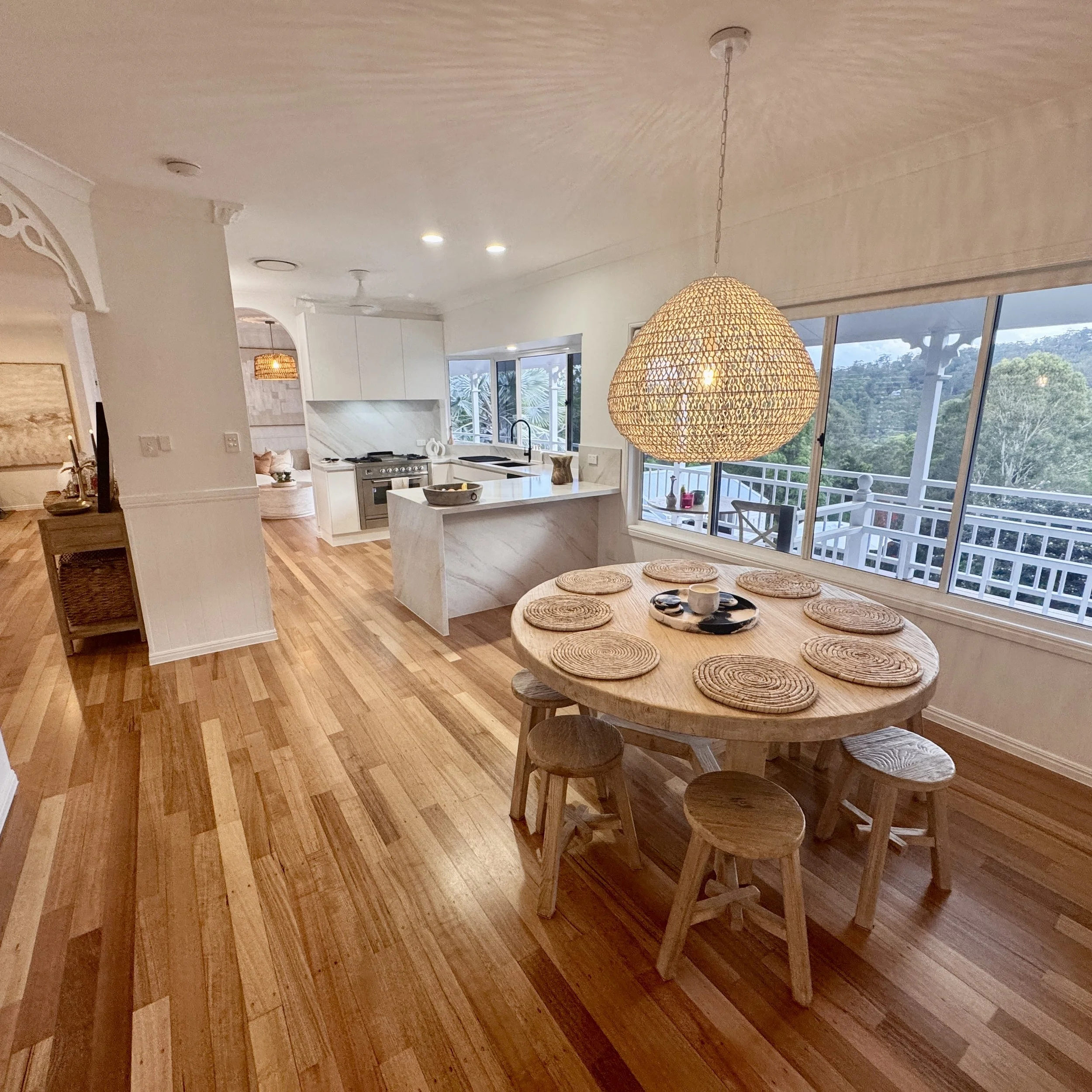 A dining area with a round wooden table and seven woven placemats, illuminated by a large woven pendant light, with large windows overlooking a balcony and greenery outside.