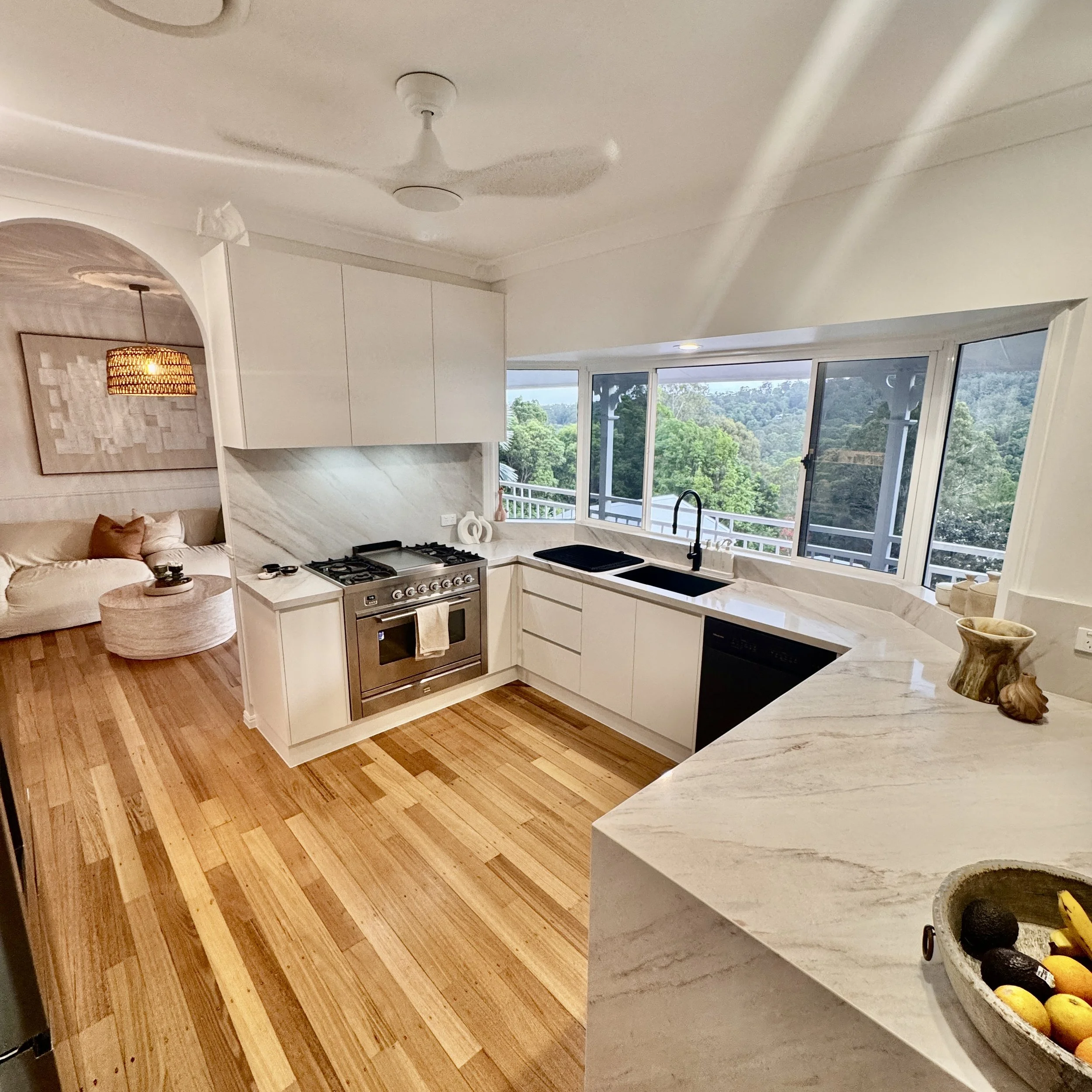 Contemporary kitchen with white cabinets, marble countertops, a black sink, stainless steel stove, and large windows overlooking a green landscape.