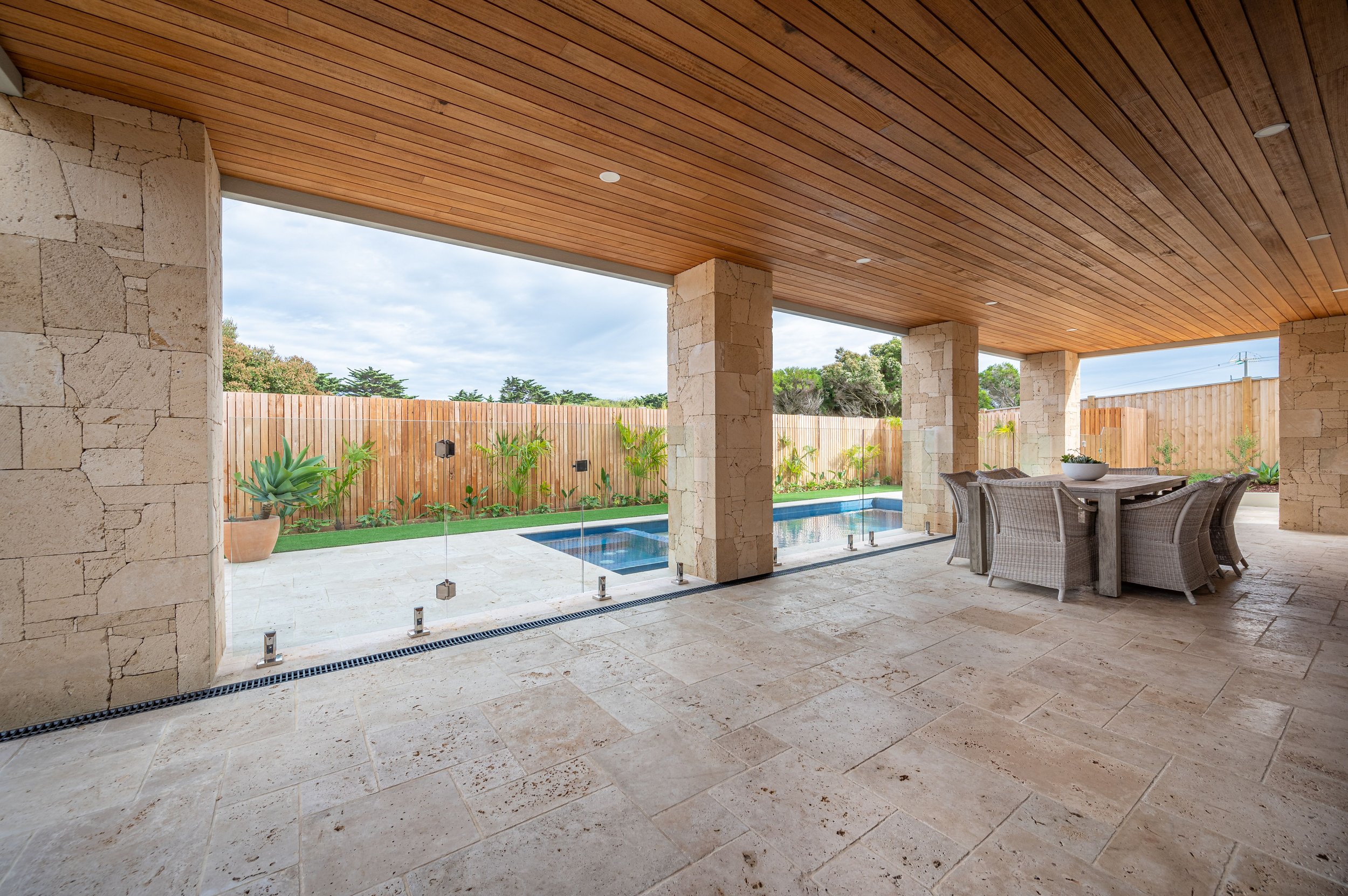 Covered outdoor patio with stone floor, wooden ceiling, glass railing, pool, greenery, and a dining table with chairs.