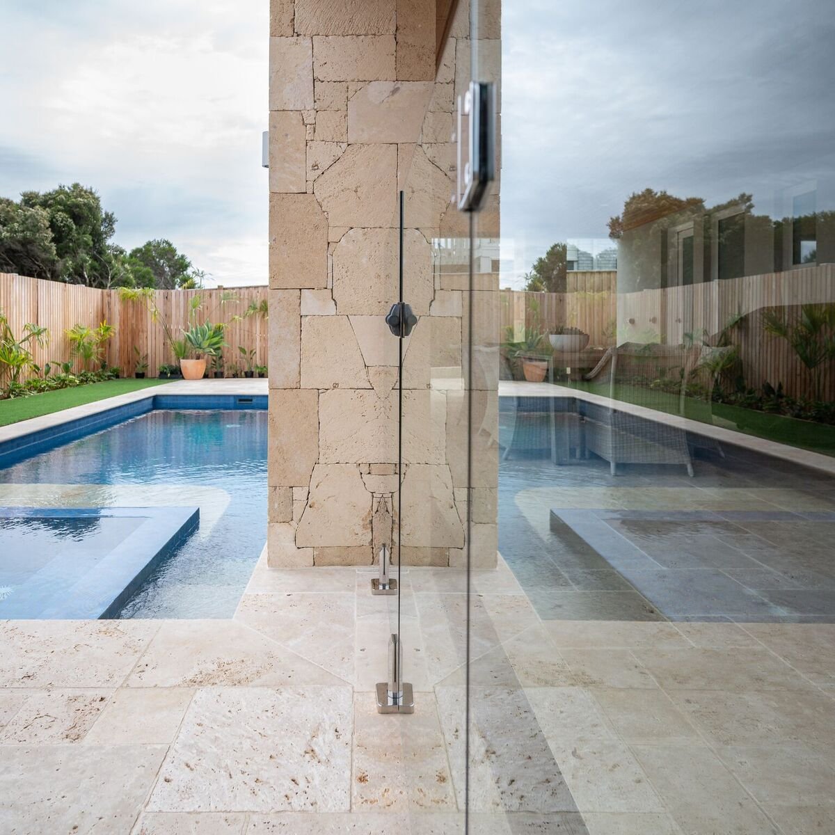 A backyard swimming pool area seen through a glass wall with a central stone pillar. The pool has a modern design with a jacuzzi section, surrounded by a wooden fence and potted plants. The sky is overcast.