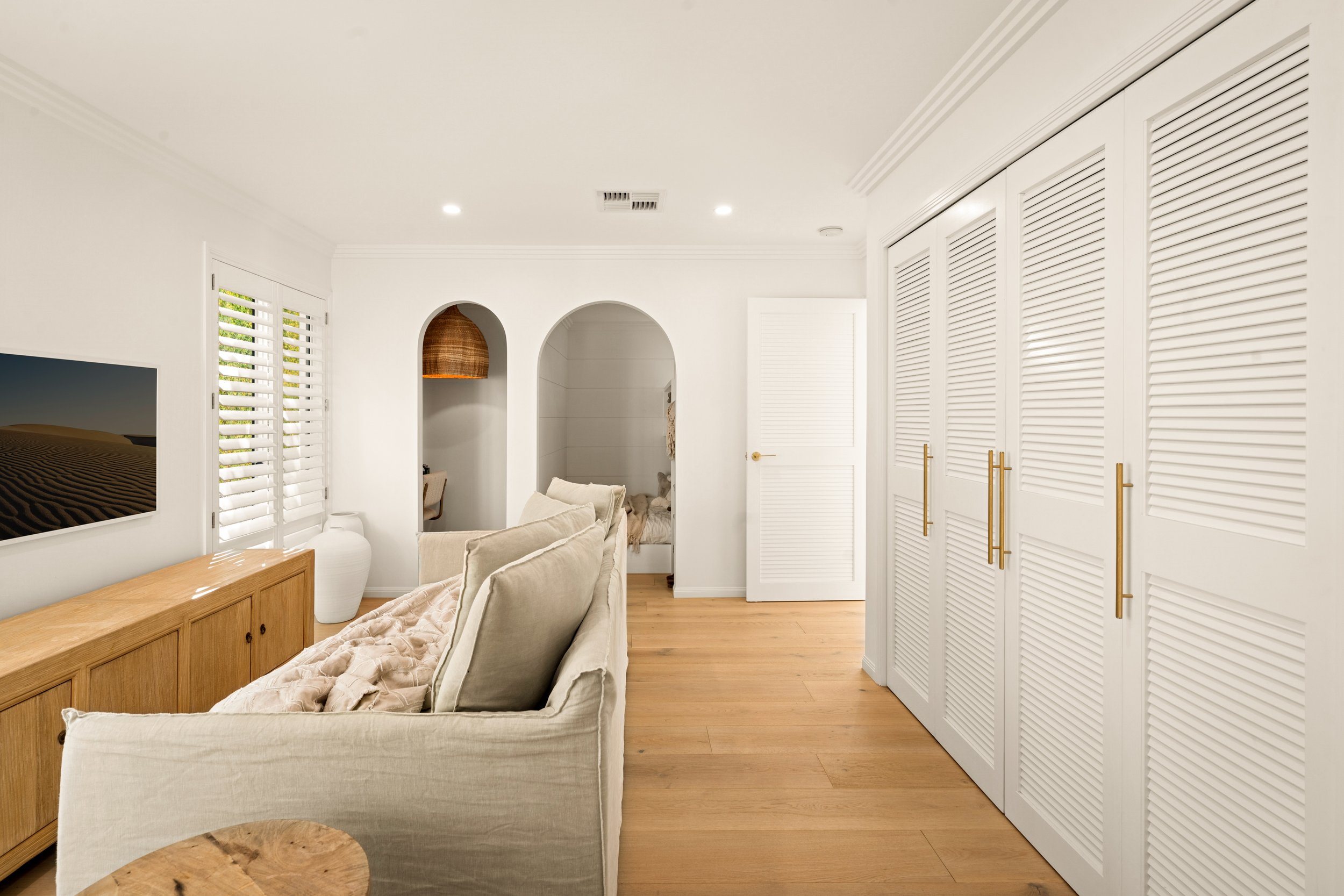 Living room with white walls, hardwood floors, a beige sofa, a wooden sideboard, and a wall-mounted television. There are windows with white shutters, a white louvered closet, and an arched doorway leading to a bedroom.