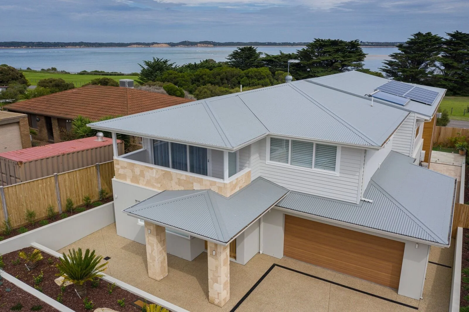 A modern two-story house with a grey metal roof, solar panels, white siding, and a beige stone accent wall, overlooking a body of water with boats in the distance.
