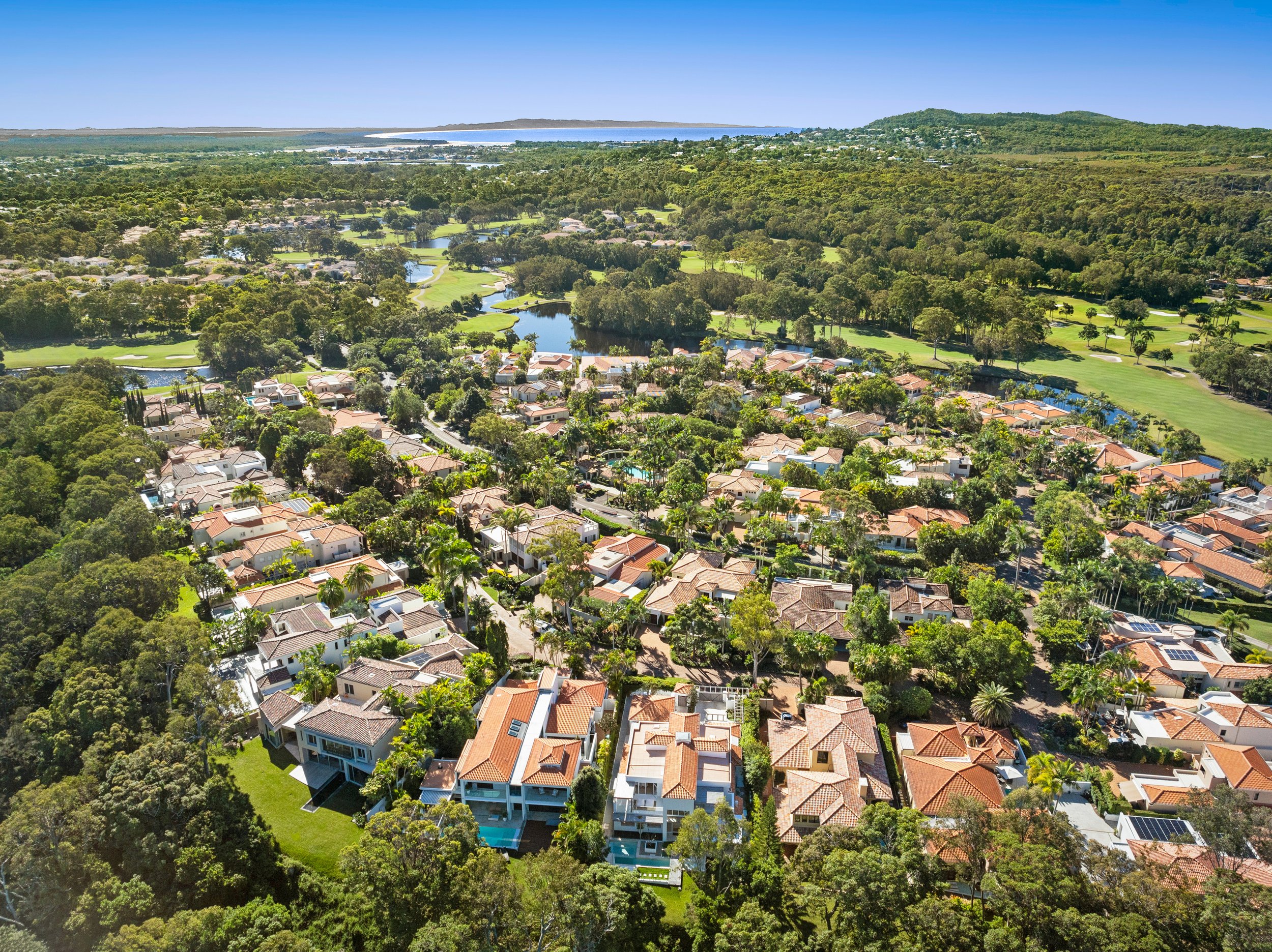 Aerial view of a suburban neighborhood with houses, trees, and green spaces, including a golf course and water bodies in the background.