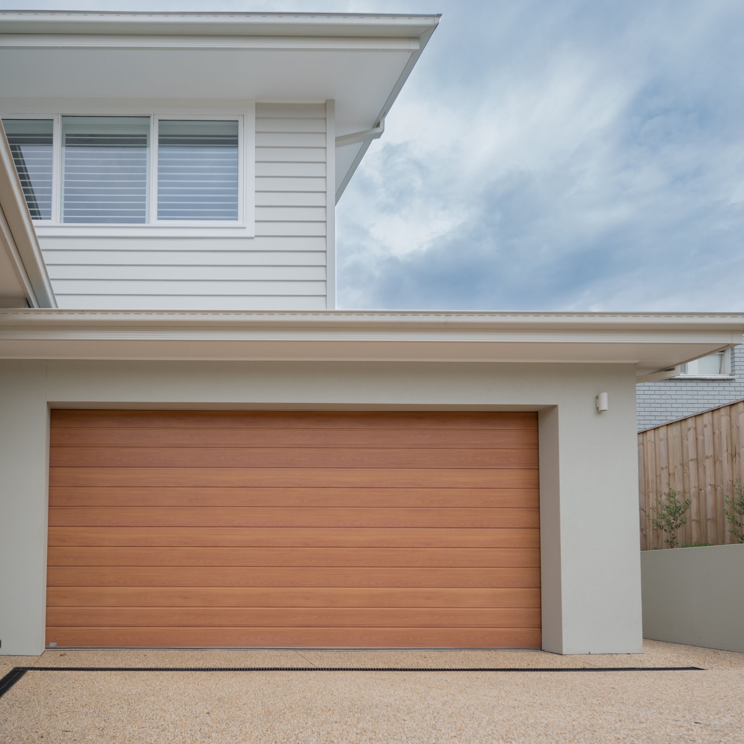 Front view of modern house with a large wooden garage door, white siding, and a window with blinds; cloudy sky in the background.