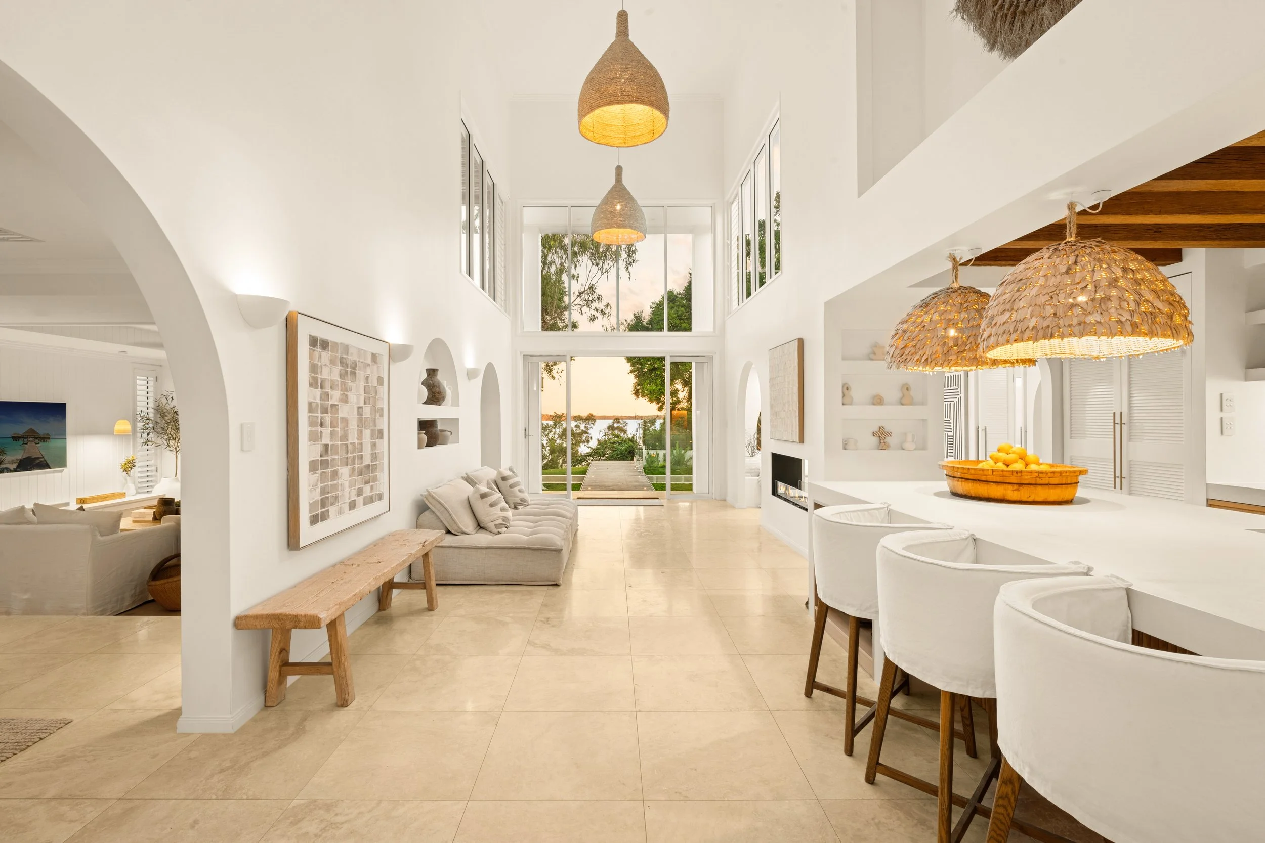 Bright, spacious living room with high ceilings, large windows, white walls, beige tile flooring, a sofa with pillows, wall art, built-in shelves, a fireplace, and a view of water and trees outside.