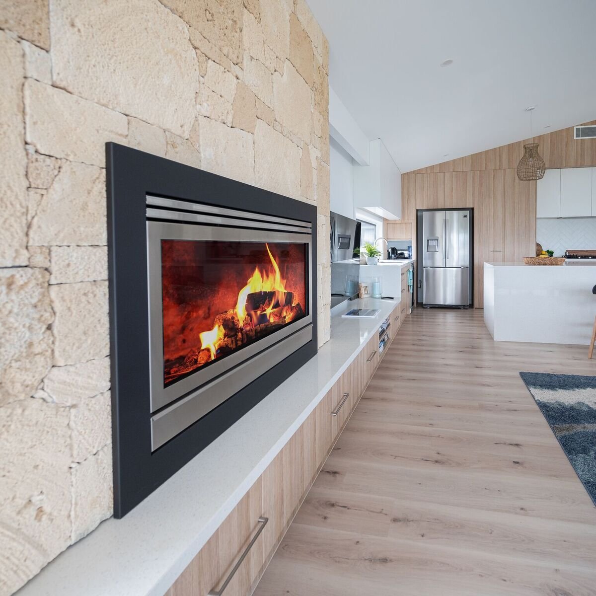 Living room with a stone fireplace, modern kitchen, stainless steel refrigerator, wooden flooring, and contemporary decor.